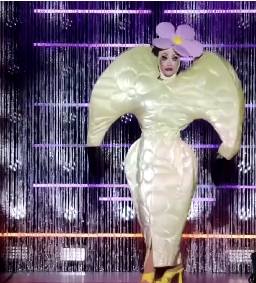 Person in avant-garde outfit with exaggerated sleeves and a large flower headpiece, standing in front of a metallic fringe backdrop