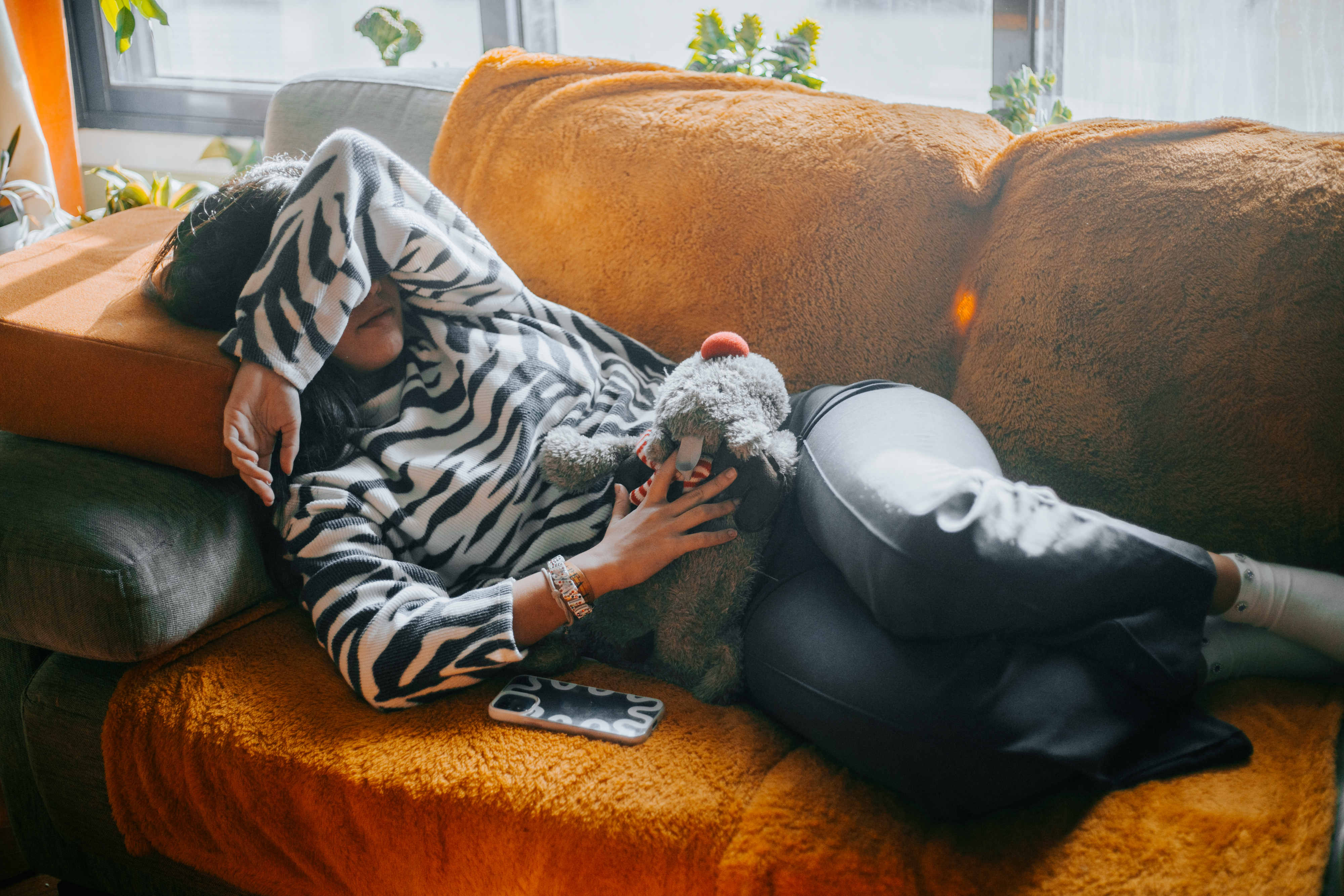 Person lying on a couch with their arm covering their face, wearing a zebra-patterned sweater, holding a stuffed toy, and a phone resting nearby