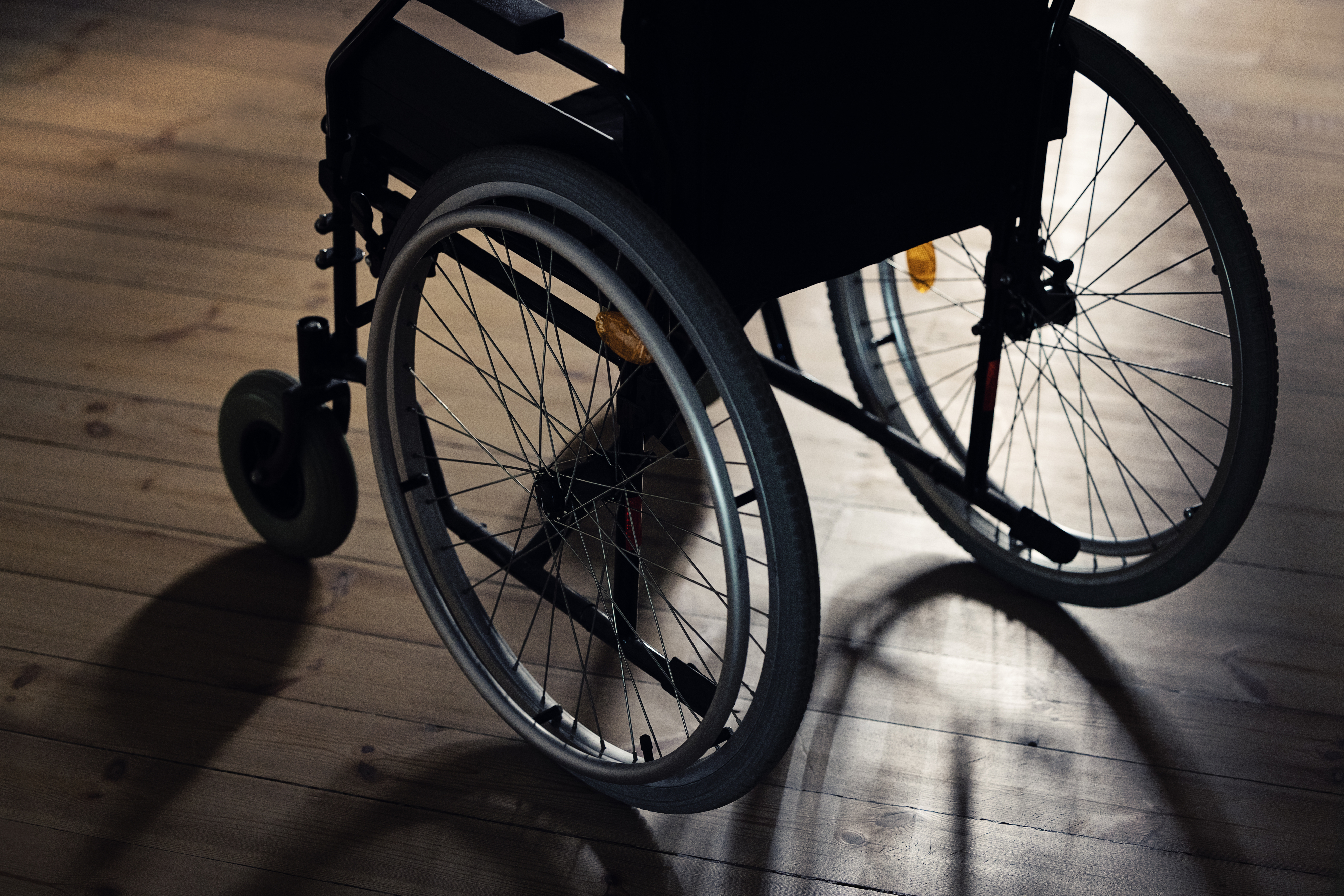 A lone wheelchair is depicted on a wooden floor, with light casting shadows through the spokes of the wheels. No people are in the image