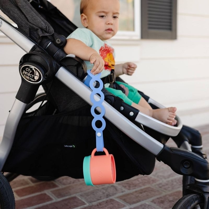 A toddler in a stroller holds a silicone baby teether with attached cups. The article is categorized as Shopping