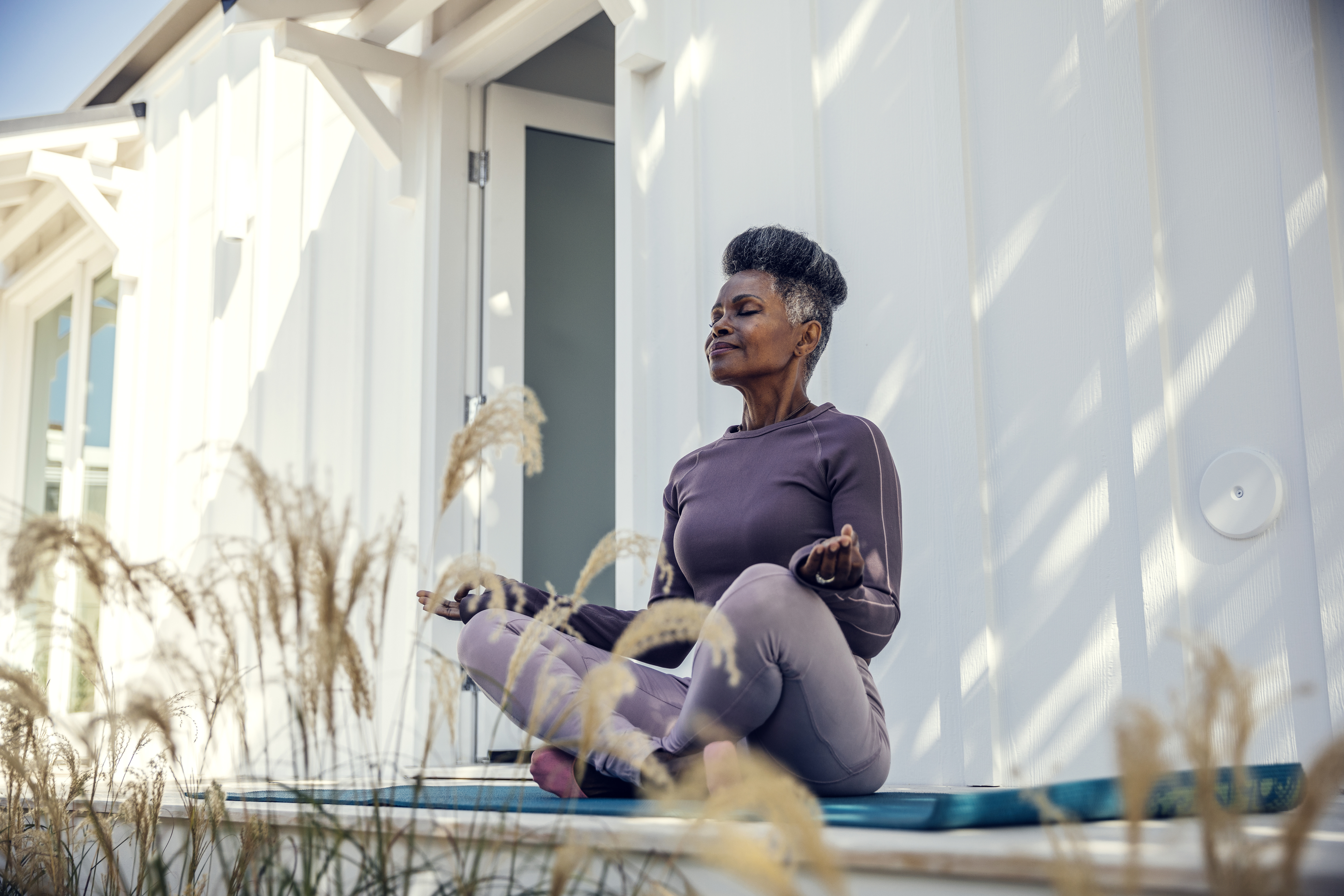 A woman in athletic wear sits cross-legged on a yoga mat with eyes closed, meditating outside a building