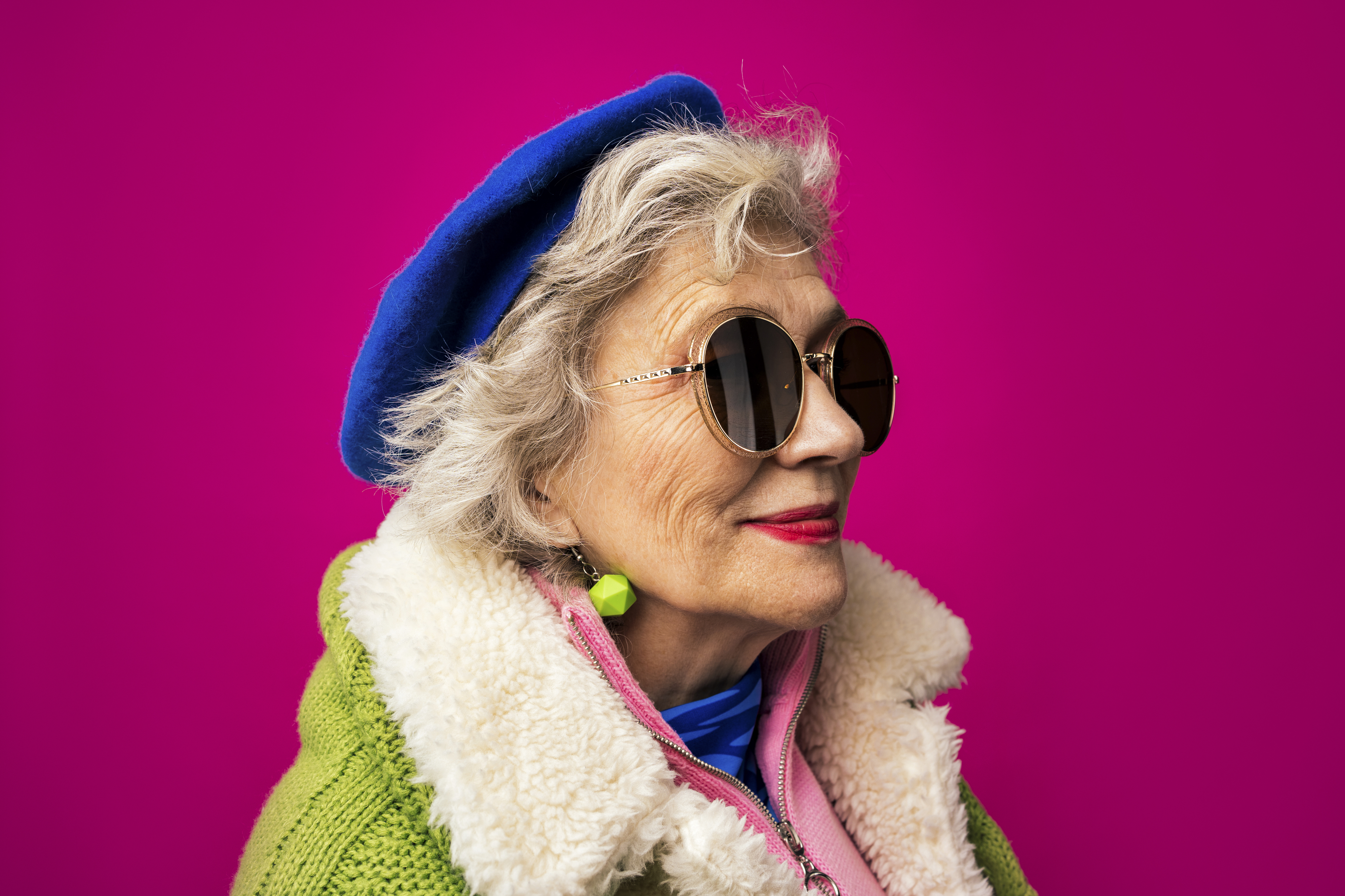 An elderly woman wearing round sunglasses, a blue beret, and a green coat with a fluffy collar stands in front of a pink background