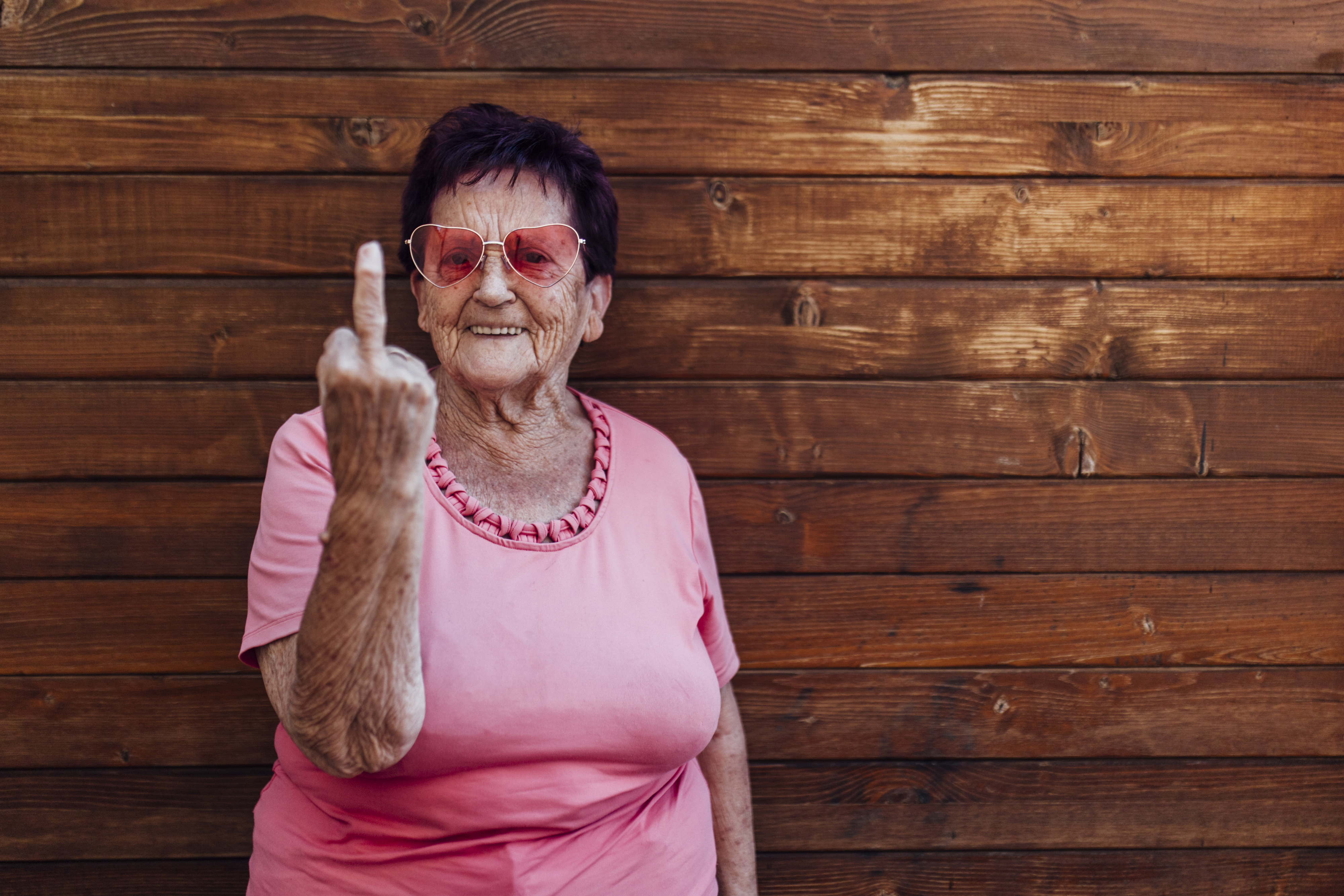 An elderly woman in a pink top and pink glasses stands against a wooden wall, smiling mischievously while raising her middle finger at the camera