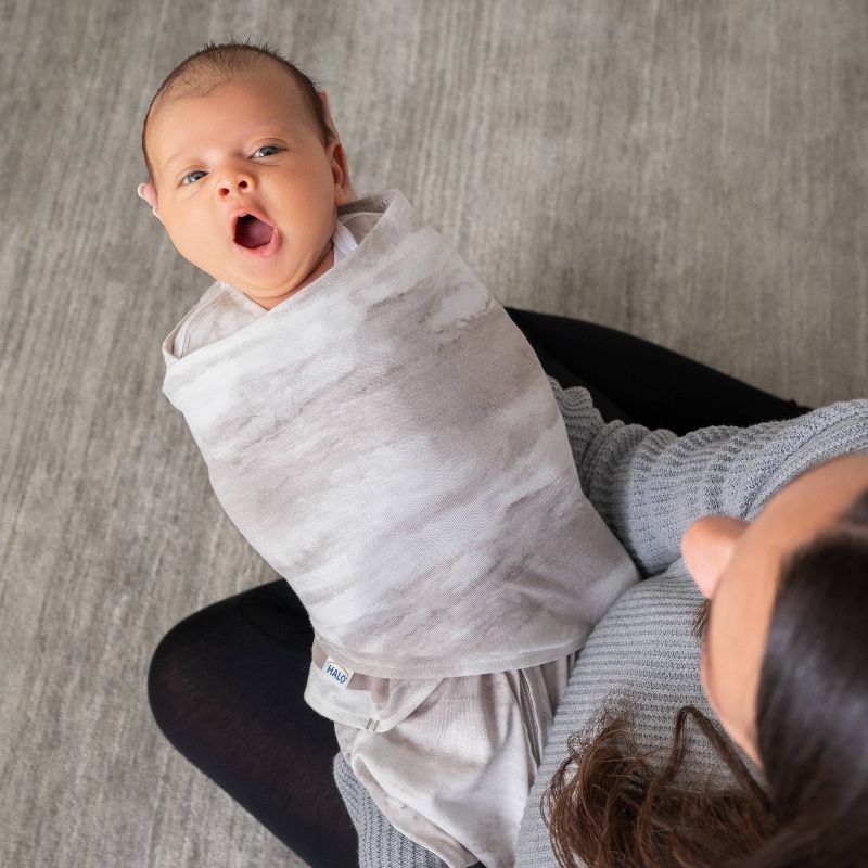 Baby wrapped in a swaddle blanket yawning while being held by an adult wearing a sweater, viewed from above