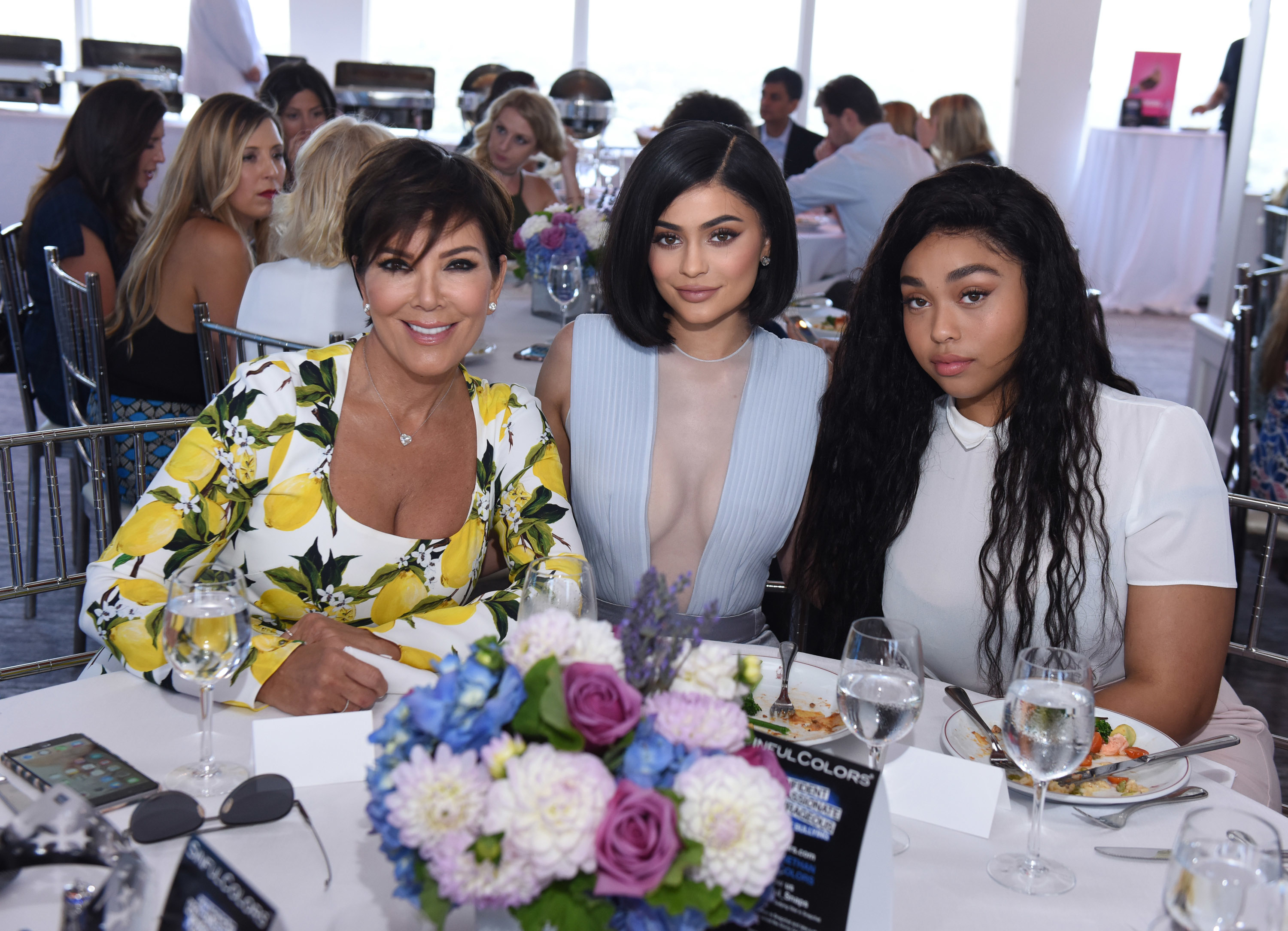 Kris Jenner, Kylie Jenner, and Jordyn Woods are seated together at an event table with flowers and food