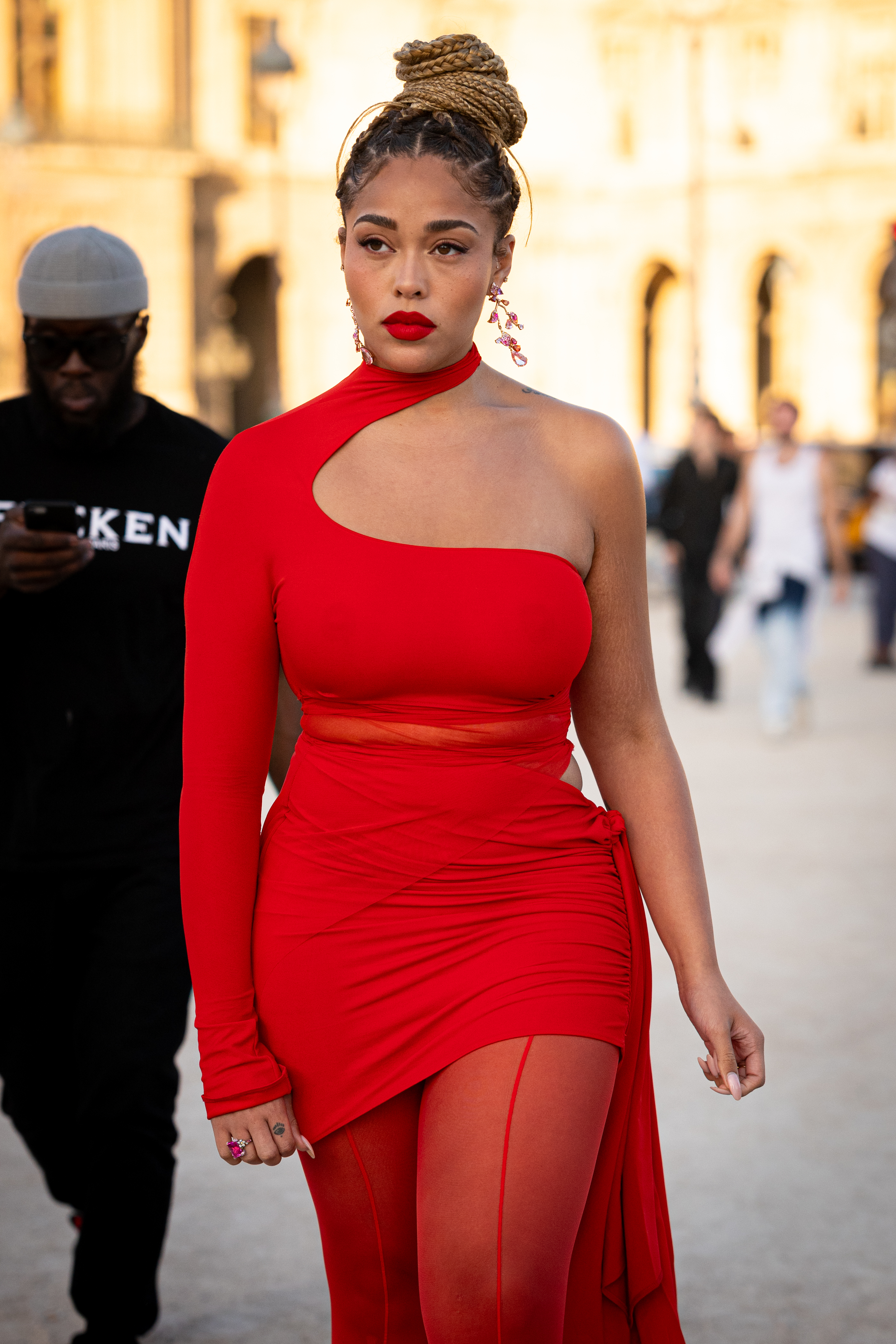 Jordyn Woods is wearing a fitted, asymmetrical dress with sheer panels, walks outdoors while a man in a hat and sunglasses is in the background