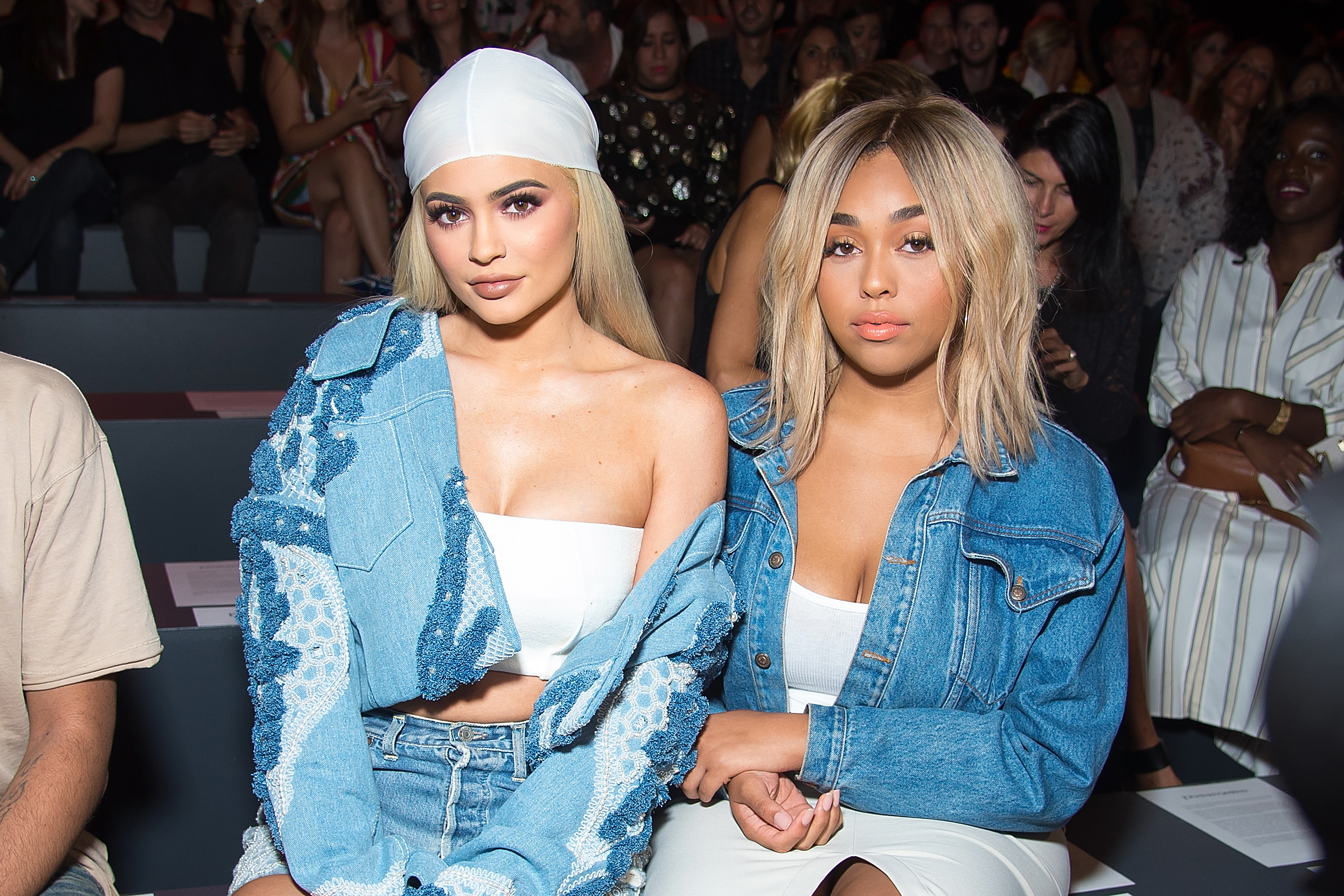 Kylie Jenner and Jordyn Woods sit front row at a fashion show, both wearing stylish denim outfits