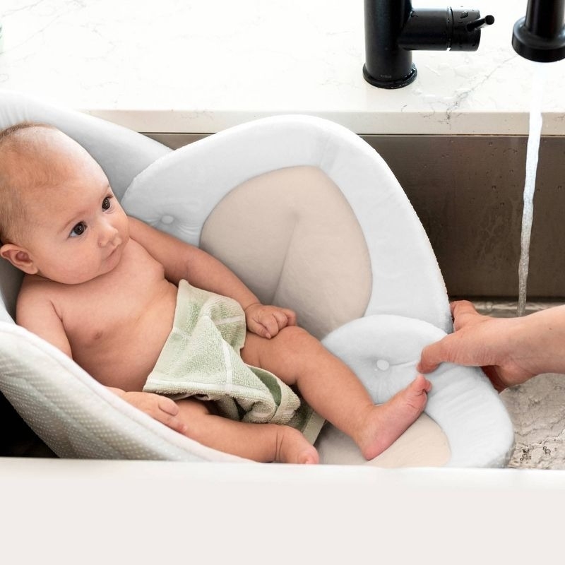 Baby sitting in a cushioned bath seat in a sink, while an adult adjusts the water. A green washcloth covers part of the baby&#x27;s torso