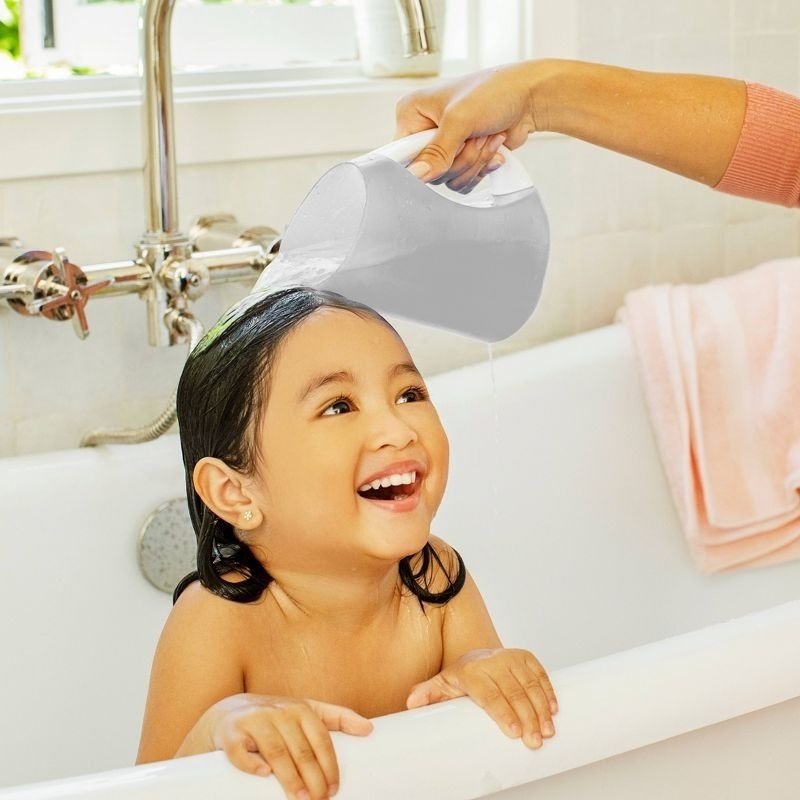 A young child smiles while being bathed in a bathtub as water is gently poured over their head by an adult hand holding a pitcher