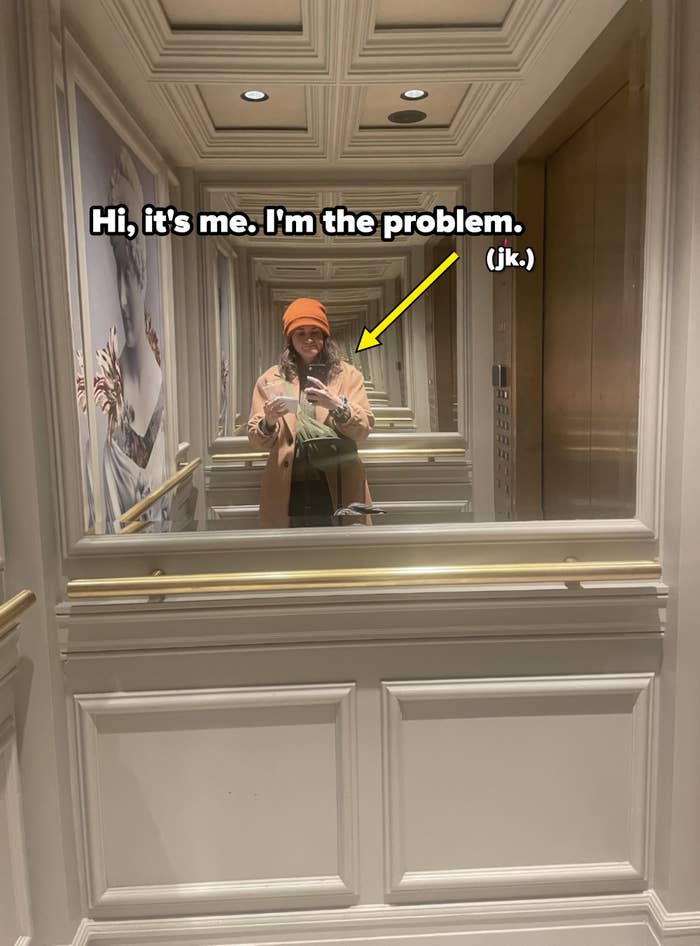 Person taking a selfie in a mirror-lined elevator, wearing a hat and casual clothing