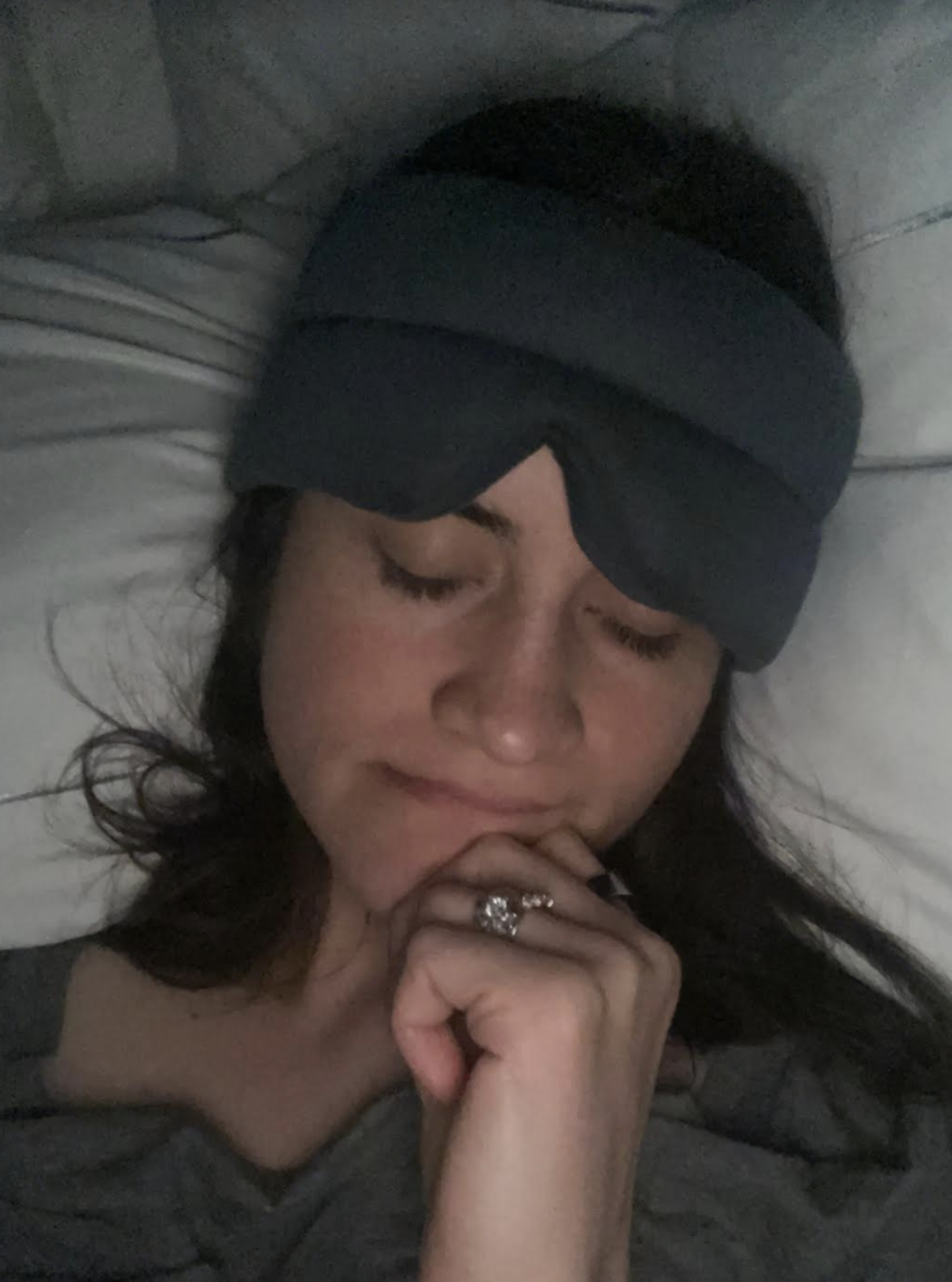 A woman wearing a sleep mask is lying in bed with her eyes closed, propped up on one hand with a thoughtful expression