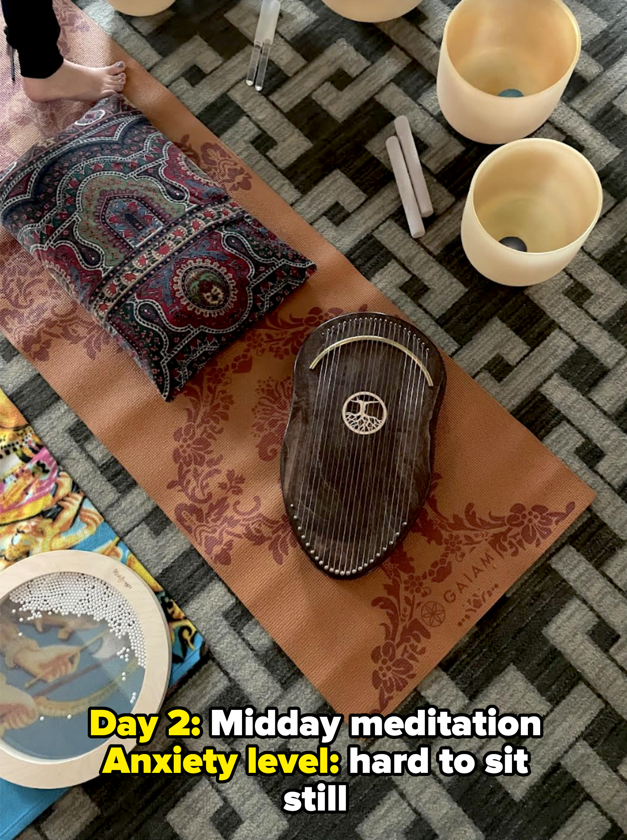 Feet standing near a patterned cushion, a musical instrument, and sound bowls on a patterned mat, indicating a meditative or sound healing setup