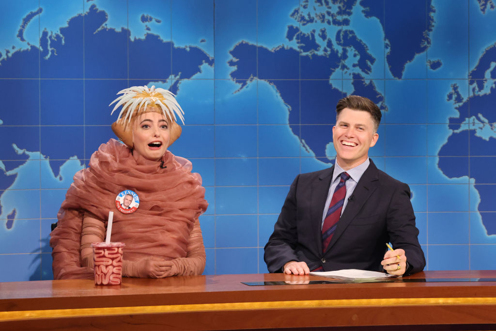 Sarah Sherman, dressed in a humorous octopus costume, sits next to Colin Jost, who is wearing a suit, on a news set with a world map backdrop