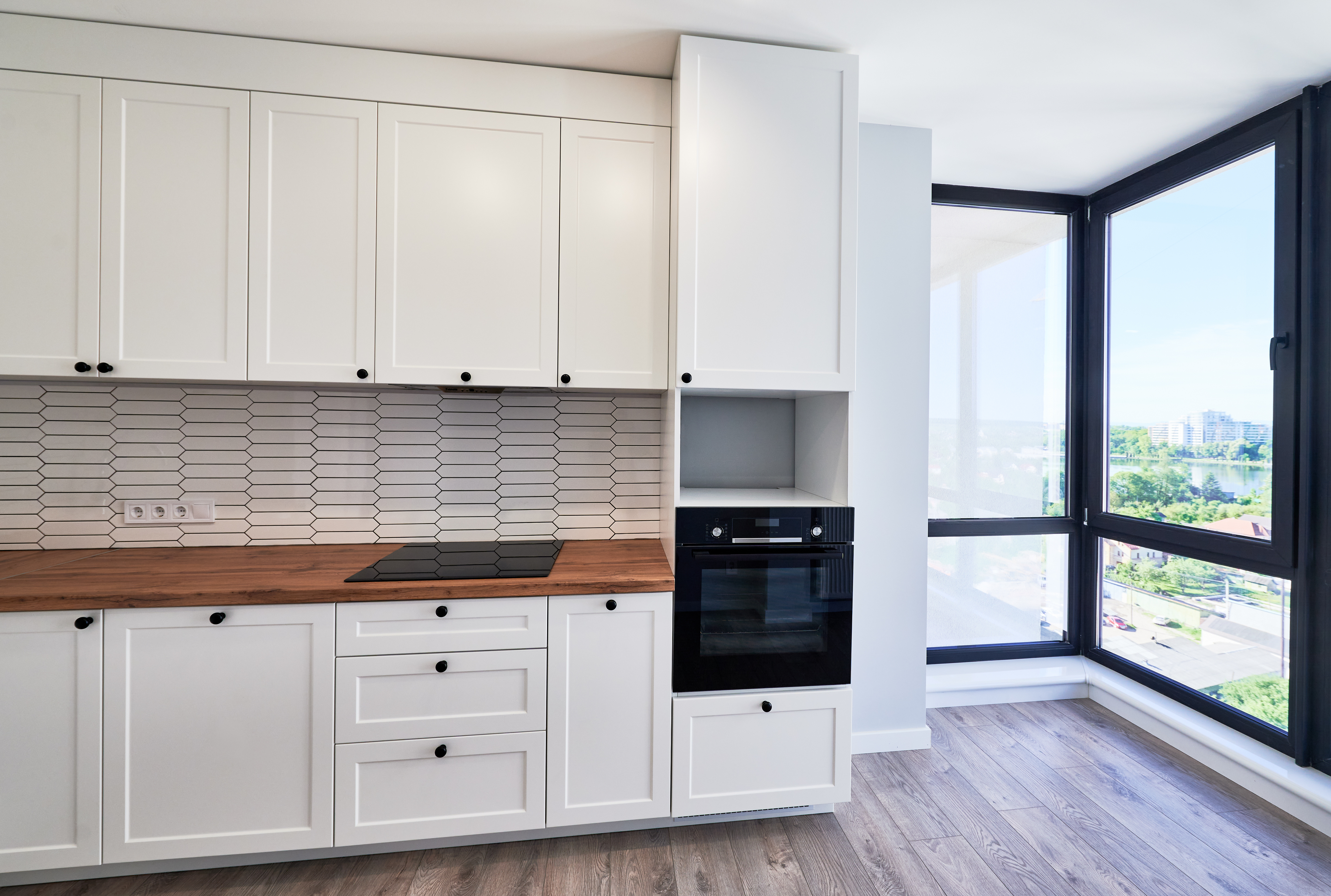 Modern kitchen with white cabinets, wood countertops, electric stovetop, built-in oven, and large corner windows showing a city view