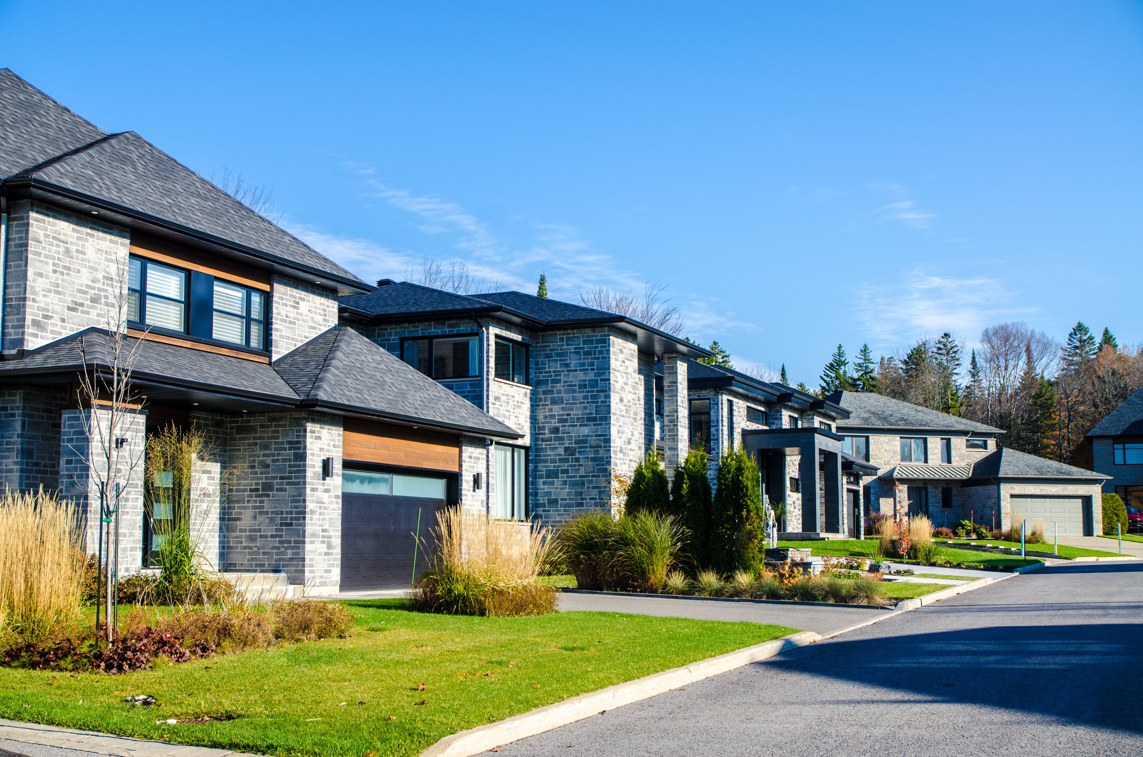 Modern suburban street lined with large, two-story houses featuring brick exteriors and well-maintained lawns. No people are visible