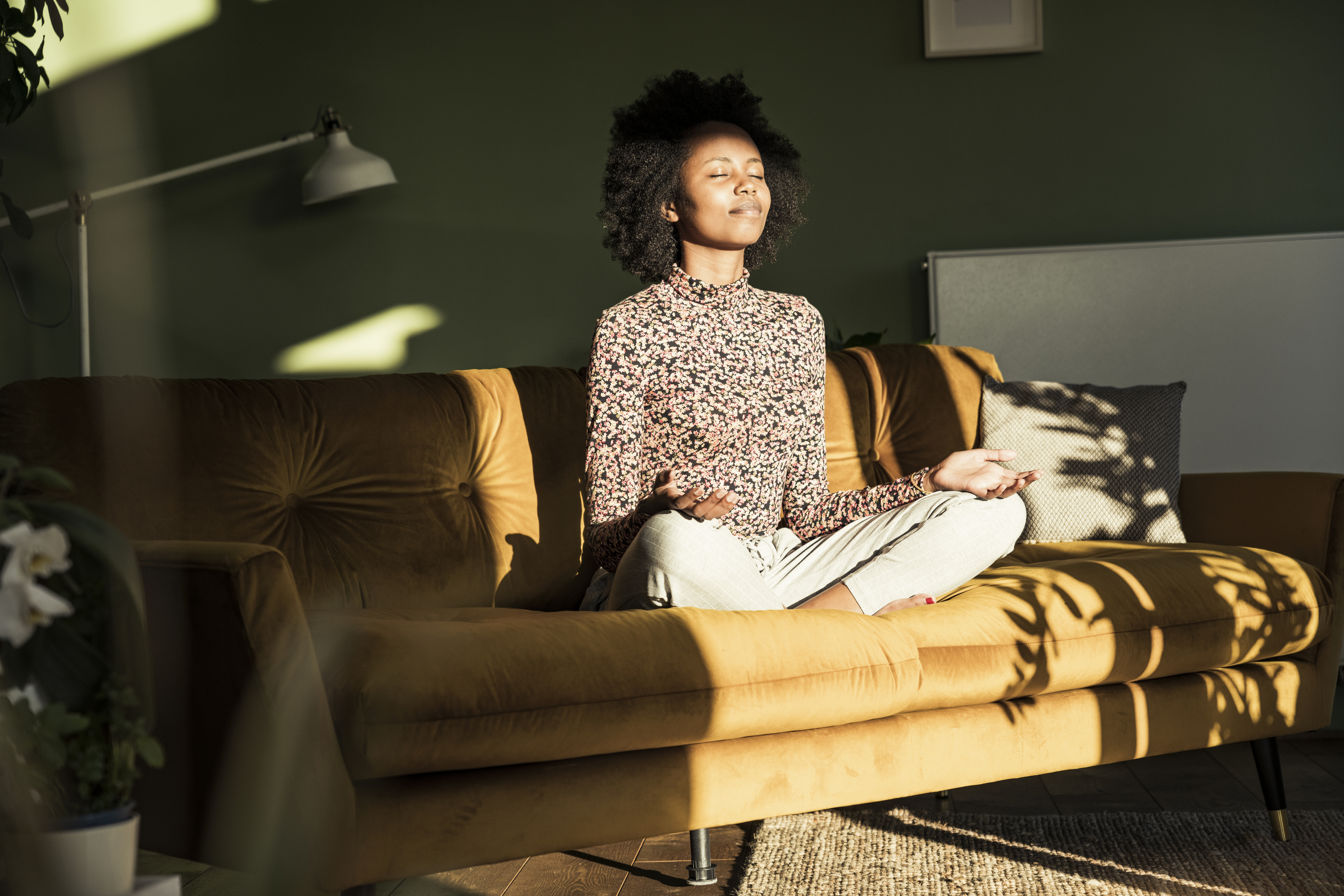 A person sits cross-legged on a couch in a sunny room, eyes closed, appearing to meditate. They are wearing a patterned top and light-colored pants