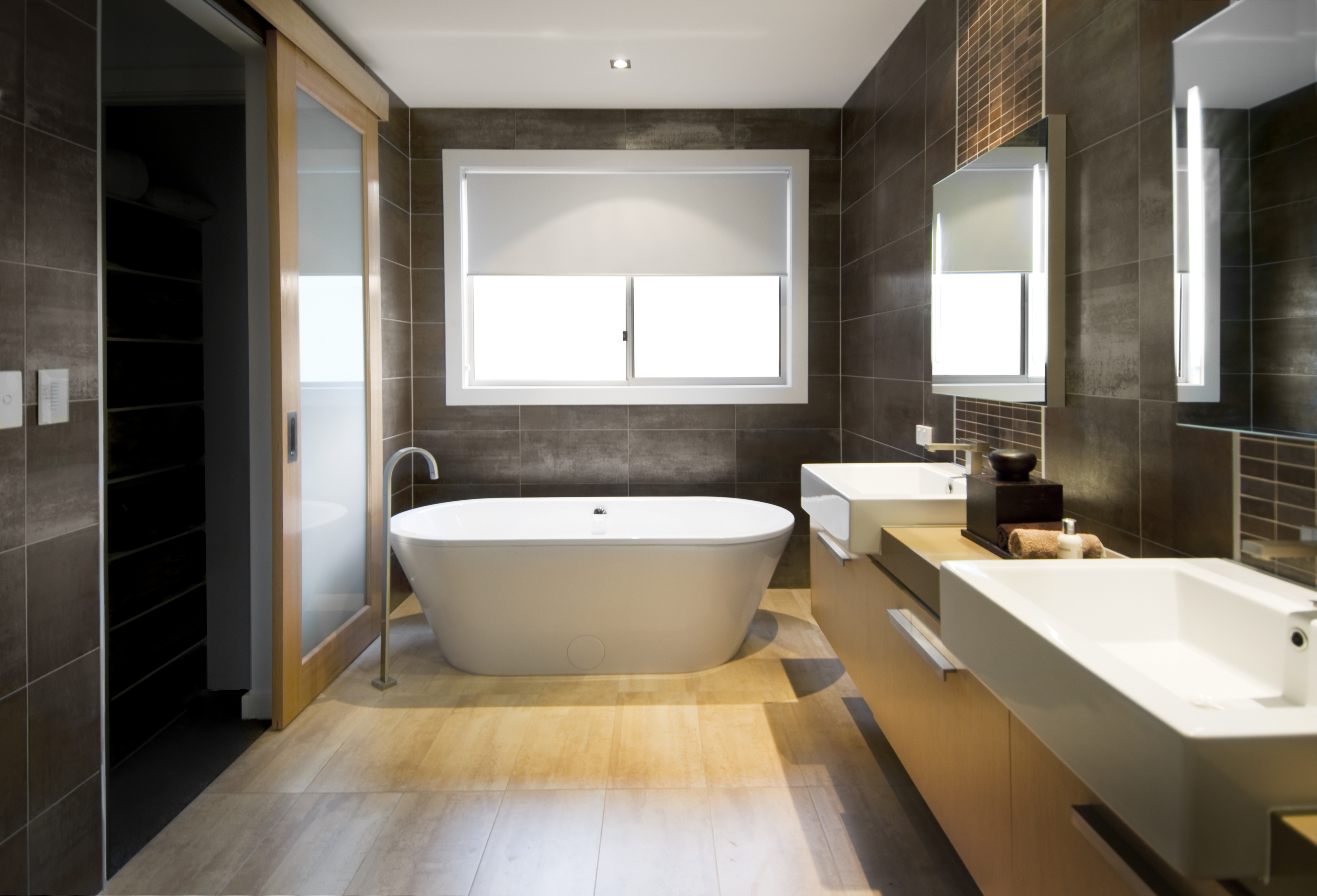 Modern bathroom with a free-standing bathtub in the center, a large window with a roller blind, and two vessel sinks on a wooden countertop