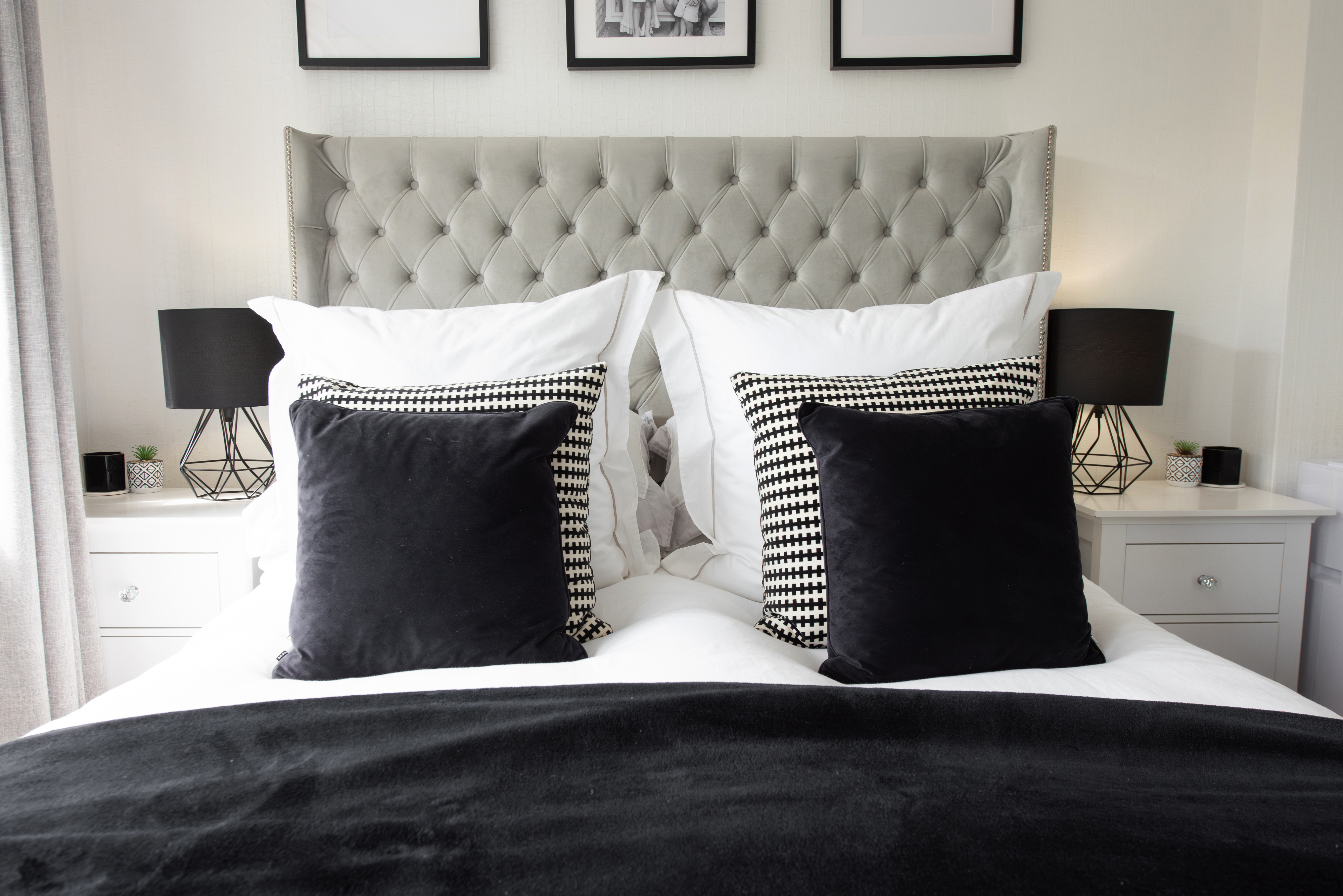 Modern bedroom with a tufted headboard, four black and white pillows, white bedding, and two nightstands with geometric lamps and small decor items