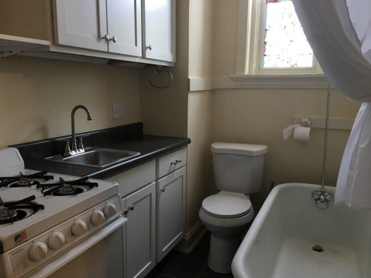 An apartment&#x27;s small kitchen with appliances beside an open bathroom with a toilet and tub, showcasing unique combined layout