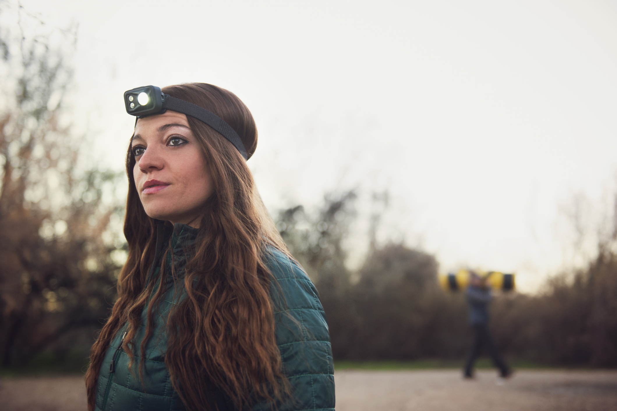 Woman wearing a headlamp and jacket stands outdoors; person with weights is blurred in the background