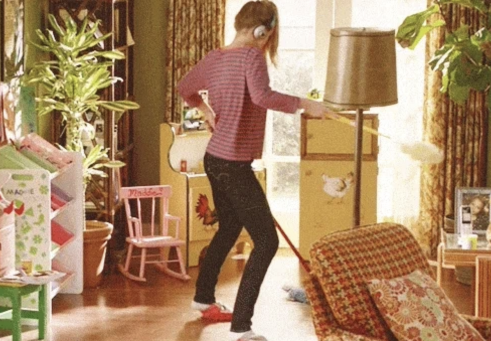 Woman wearing headphones dusting, mopping and dancing in a living room filled with plants, furniture, and children&#x27;s toys