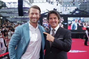 Glen Powell in a blue suit and Tom Cruise in a black suit pose and smile for photos on a red carpet at an event