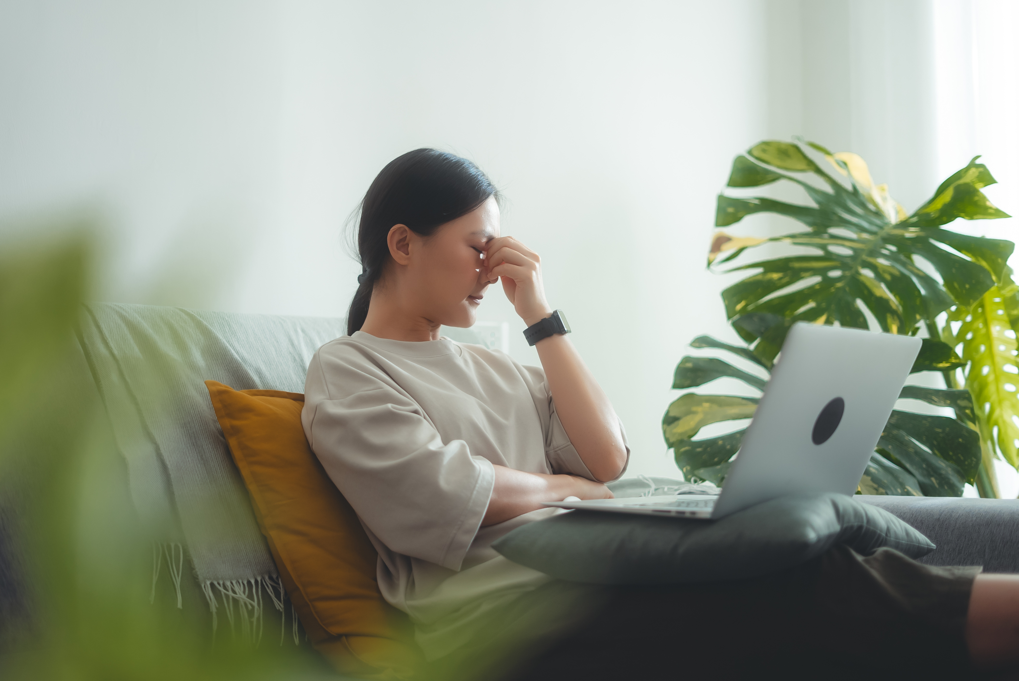 A person sits on a couch with their hand on their face, appearing stressed. They have a laptop open on their lap and a large plant is in the background