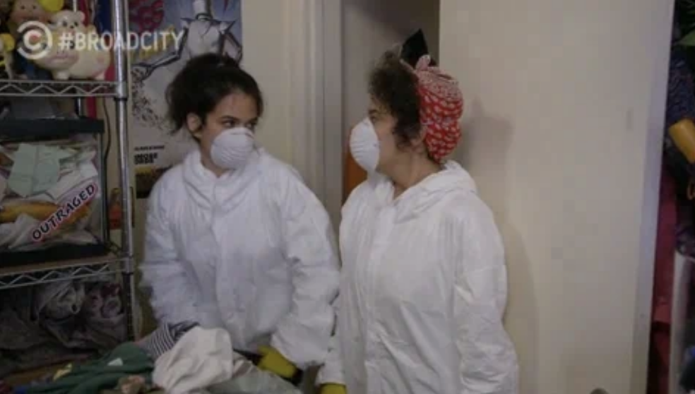 Broad City&#x27;s Abbi Jacobson and Ilana Glazer wear protective suits and face masks, standing in a cluttered room