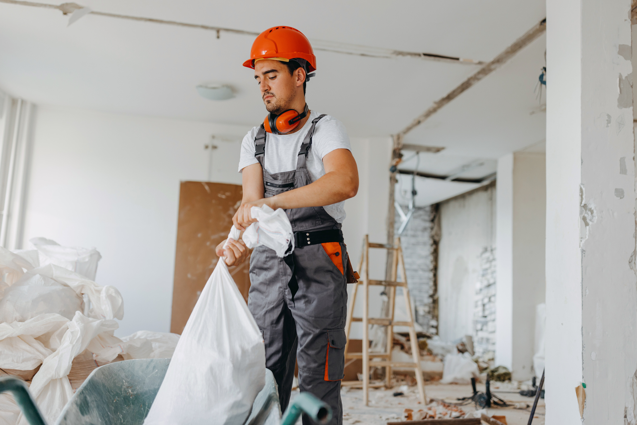 A man in a construction outfit and hard hat is working indoors, holding a large trash bag. There is construction debris and tools around him