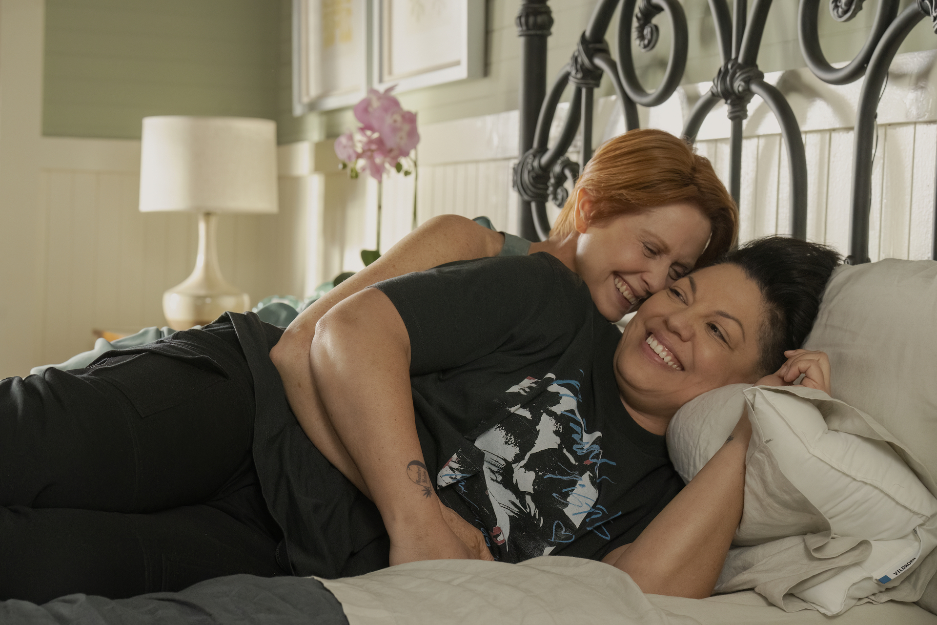 Cynthia Nixon and Sara Ramirez are lounging and smiling together on a bed in a cozy, well-decorated room