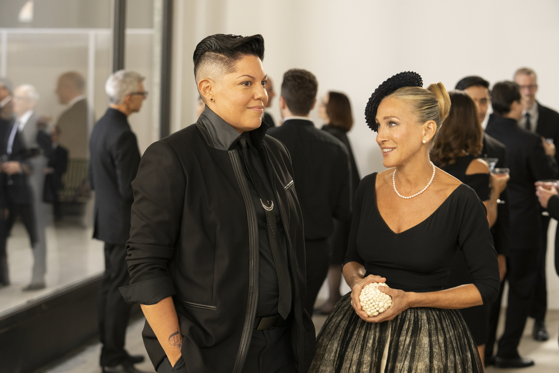 Sara Ramirez in a black suit and Sarah Jessica Parker in a dress and pearl necklace, holding a pearl clutch, at an indoor event