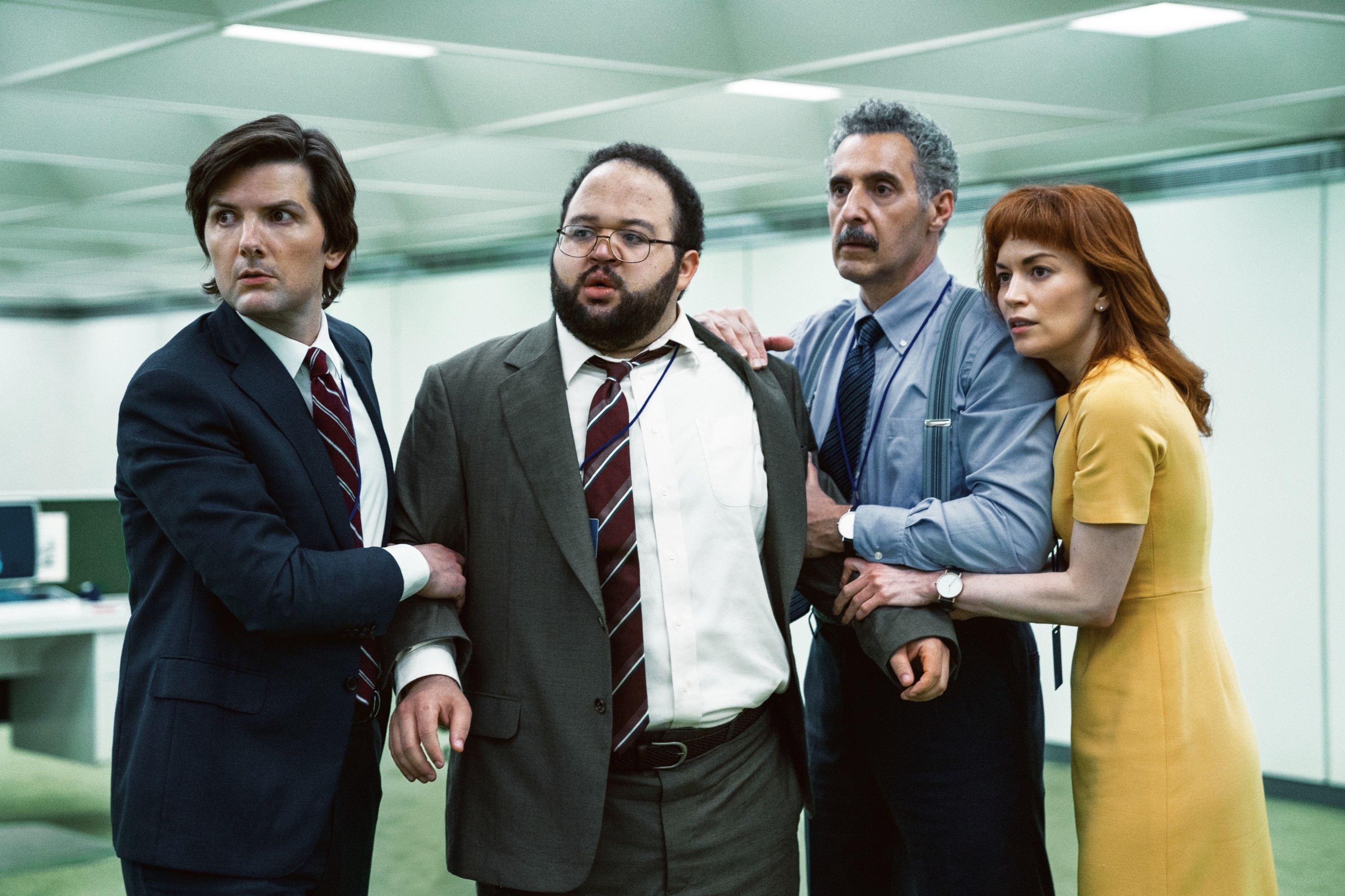 Adam Scott, Zach Cherry, John Turturro, and Britt Lower, in business attire, surround Zach in an office setting, appearing concerned