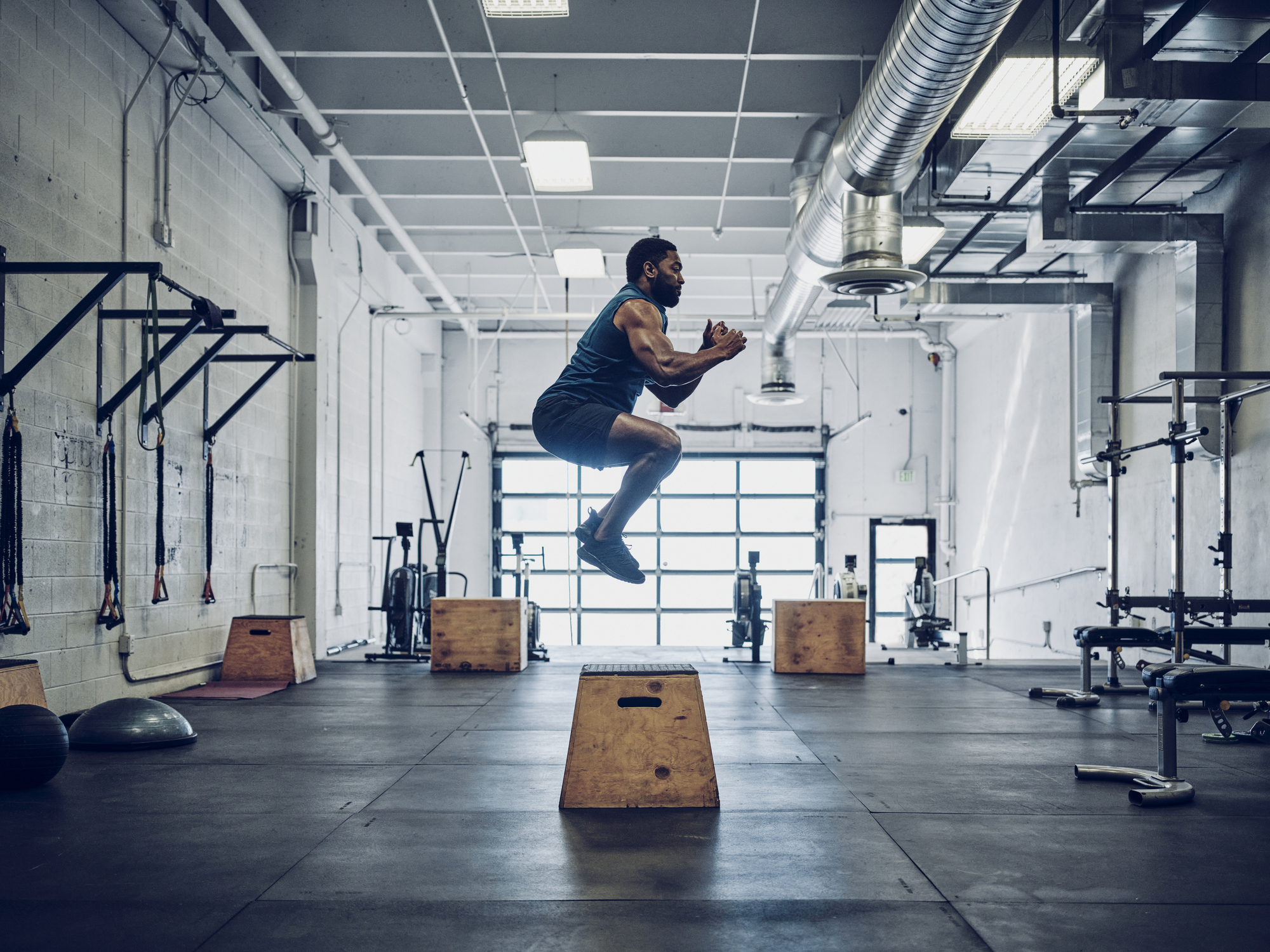 A man doing a box jump exercise in a gym. He appears focused and energetic, wearing athletic clothing. The gym is spacious with various exercise equipment