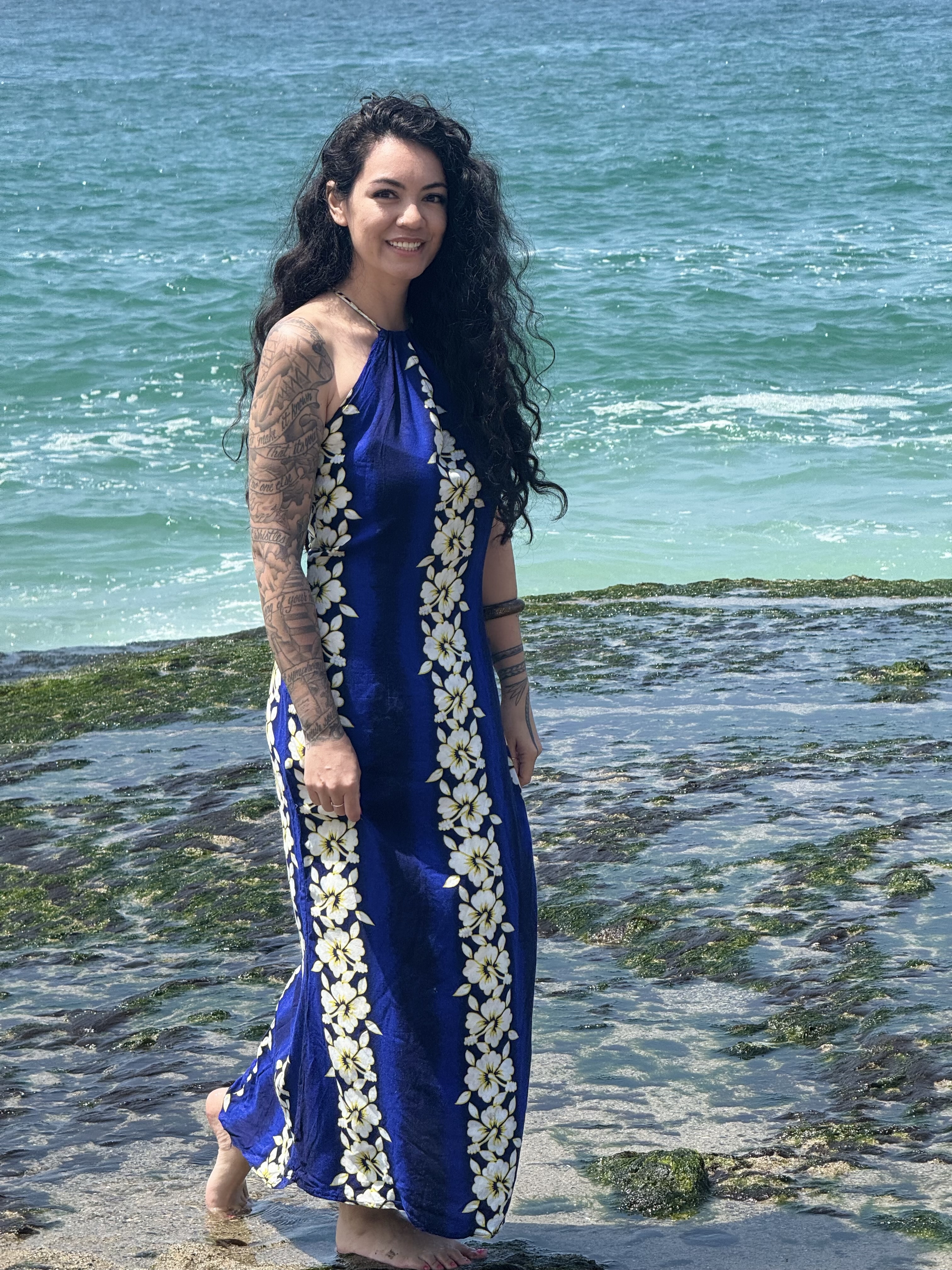 A woman with long, curly hair and tattoos on her arms is standing on a rocky beach by the ocean, wearing a floral-patterned dress