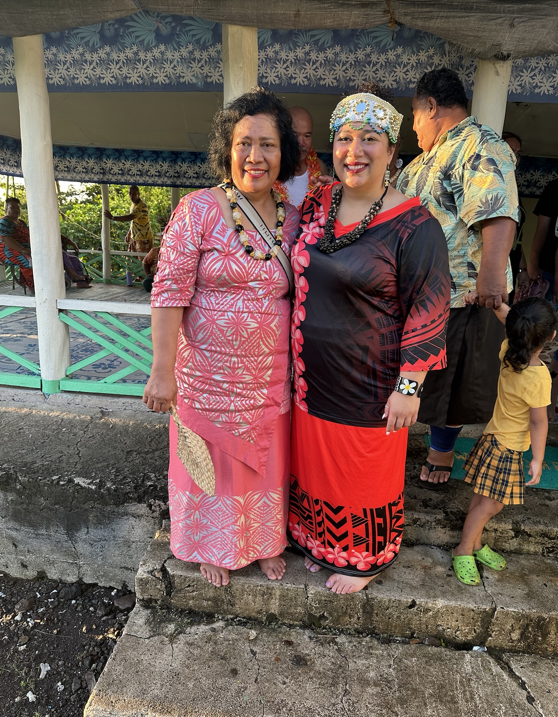 Two smiling women, dressed in traditional Pacific Islander attire, stand barefoot on stone steps. People and a child are in the background