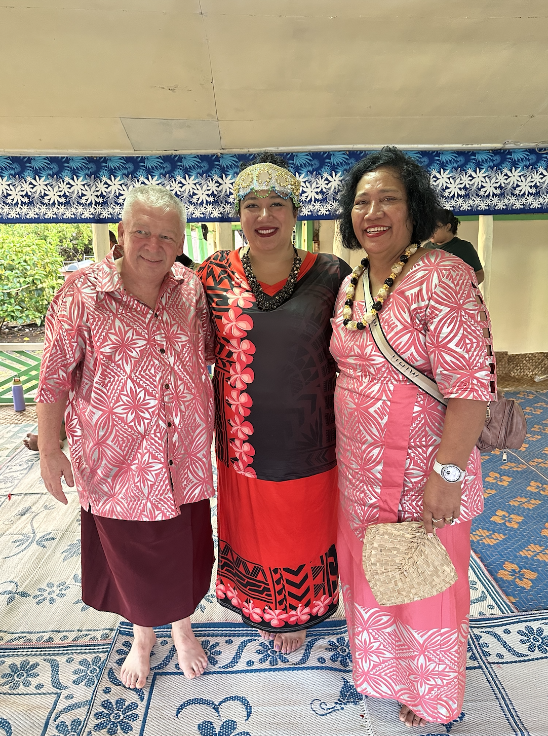 Three people in traditional Polynesian attire are smiling and standing together in a room with patterned decor
