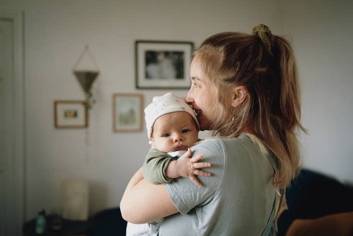 A woman holds a baby close while smiling in a cozy room with framed pictures on the wall