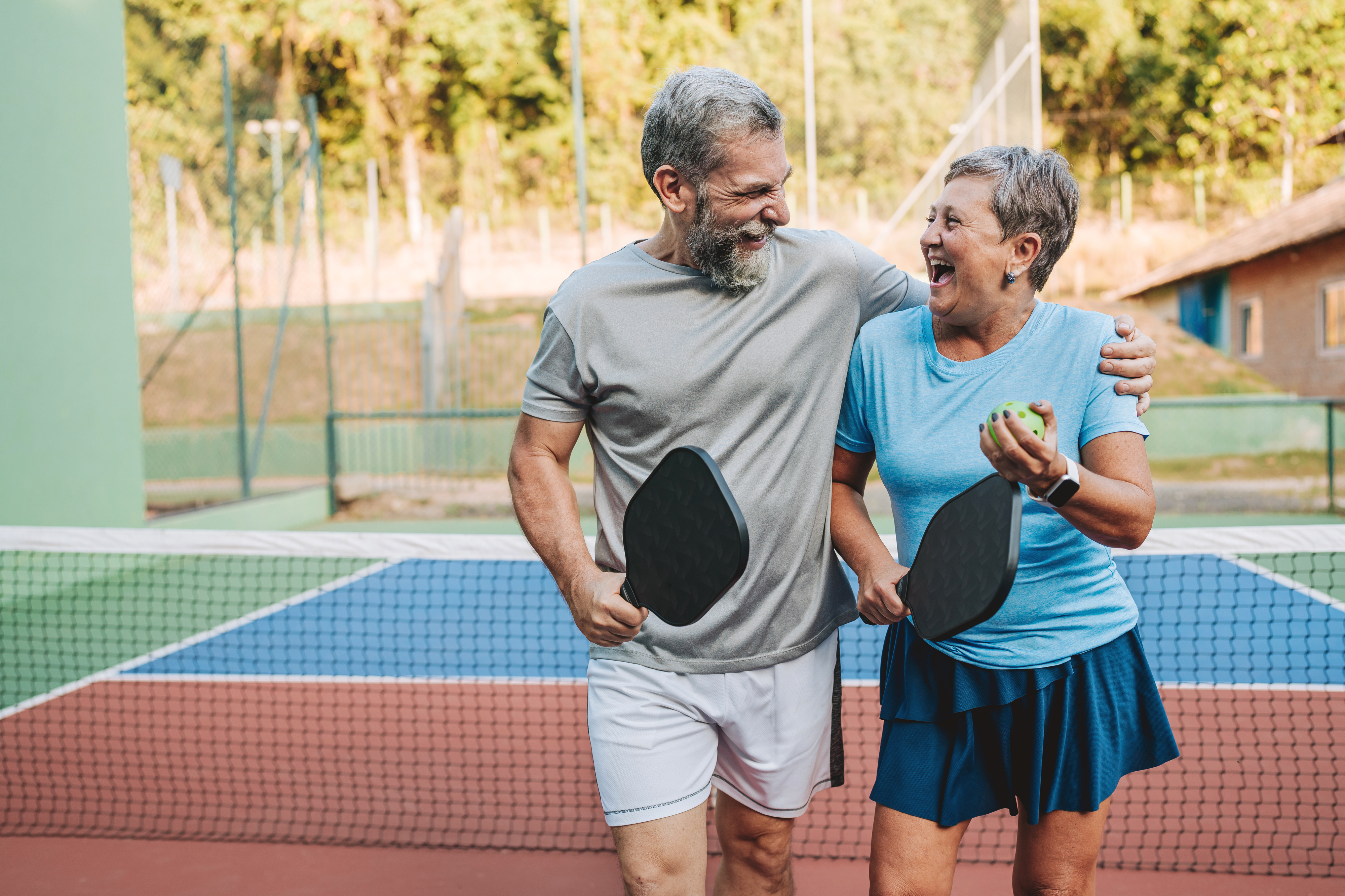 An elderly man and woman in sporty outfits smiling and holding pickleball paddles on a court. The woman also holds a pickleball