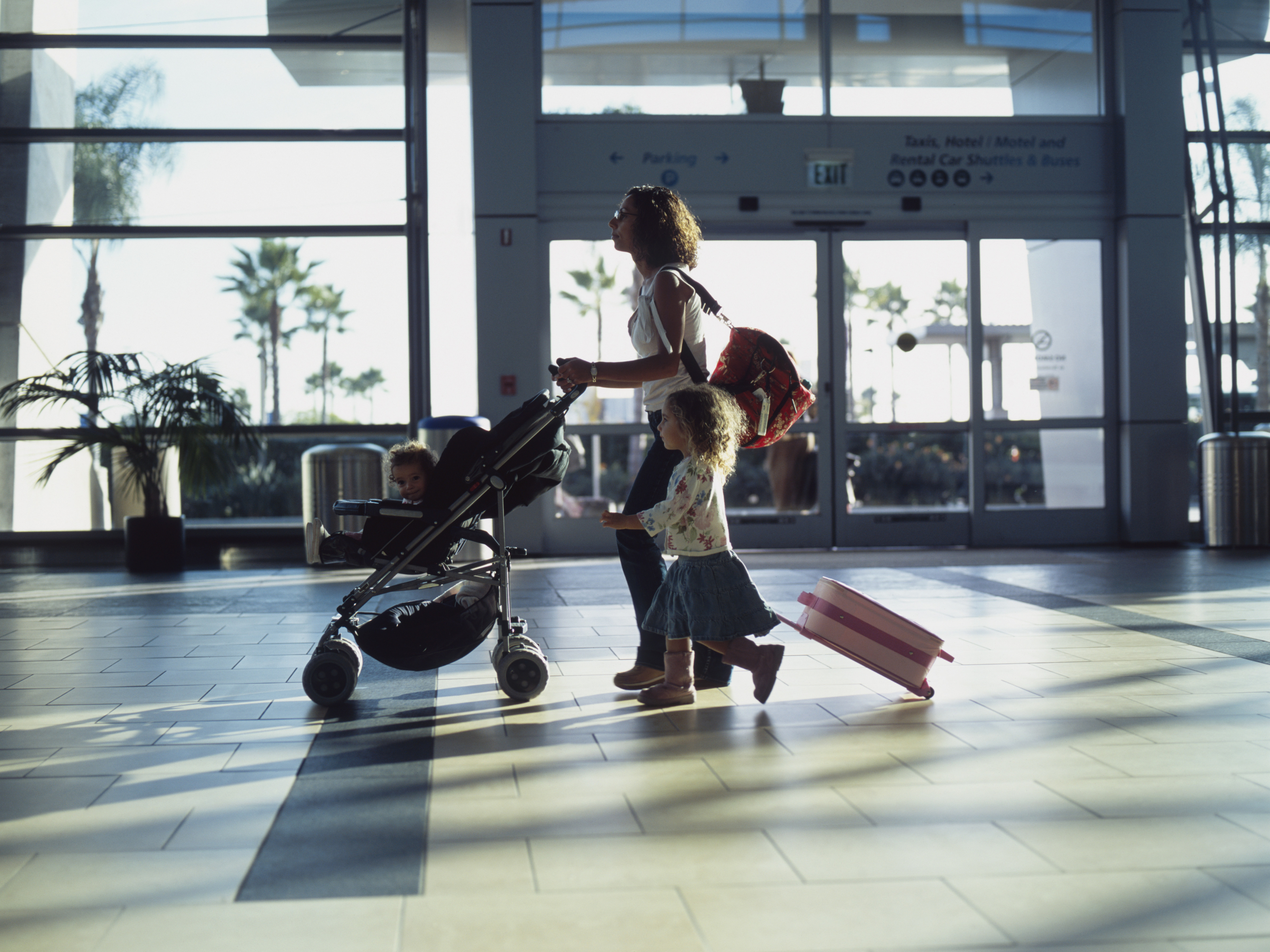A woman pushes a stroller with a child and walks beside another child pulling a suitcase through an airport terminal