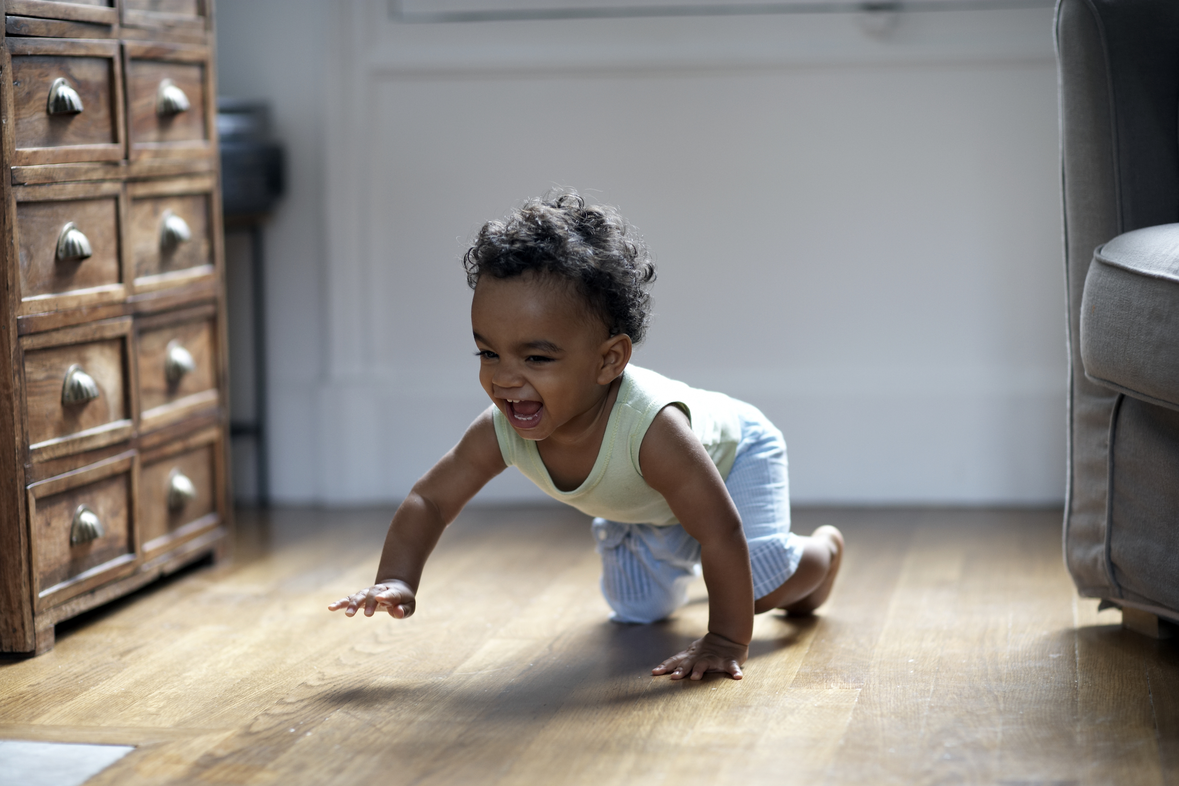 A joyful baby crawls on a wooden floor in a well-lit room, smiling and reaching forward