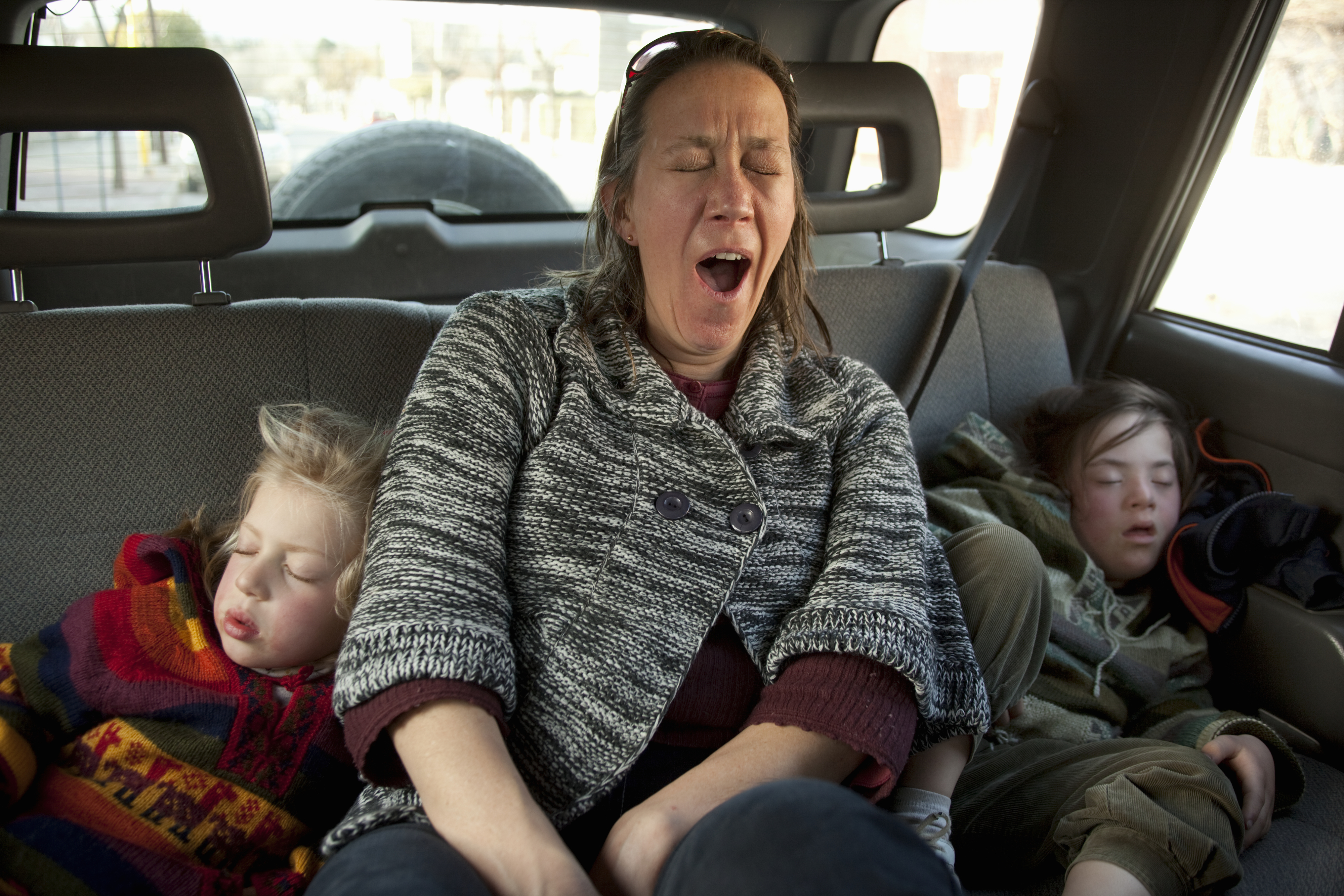 A woman yawns while sitting in a car&#x27;s backseat with two sleeping children beside her. The children are dressed in colorful and cozy clothing