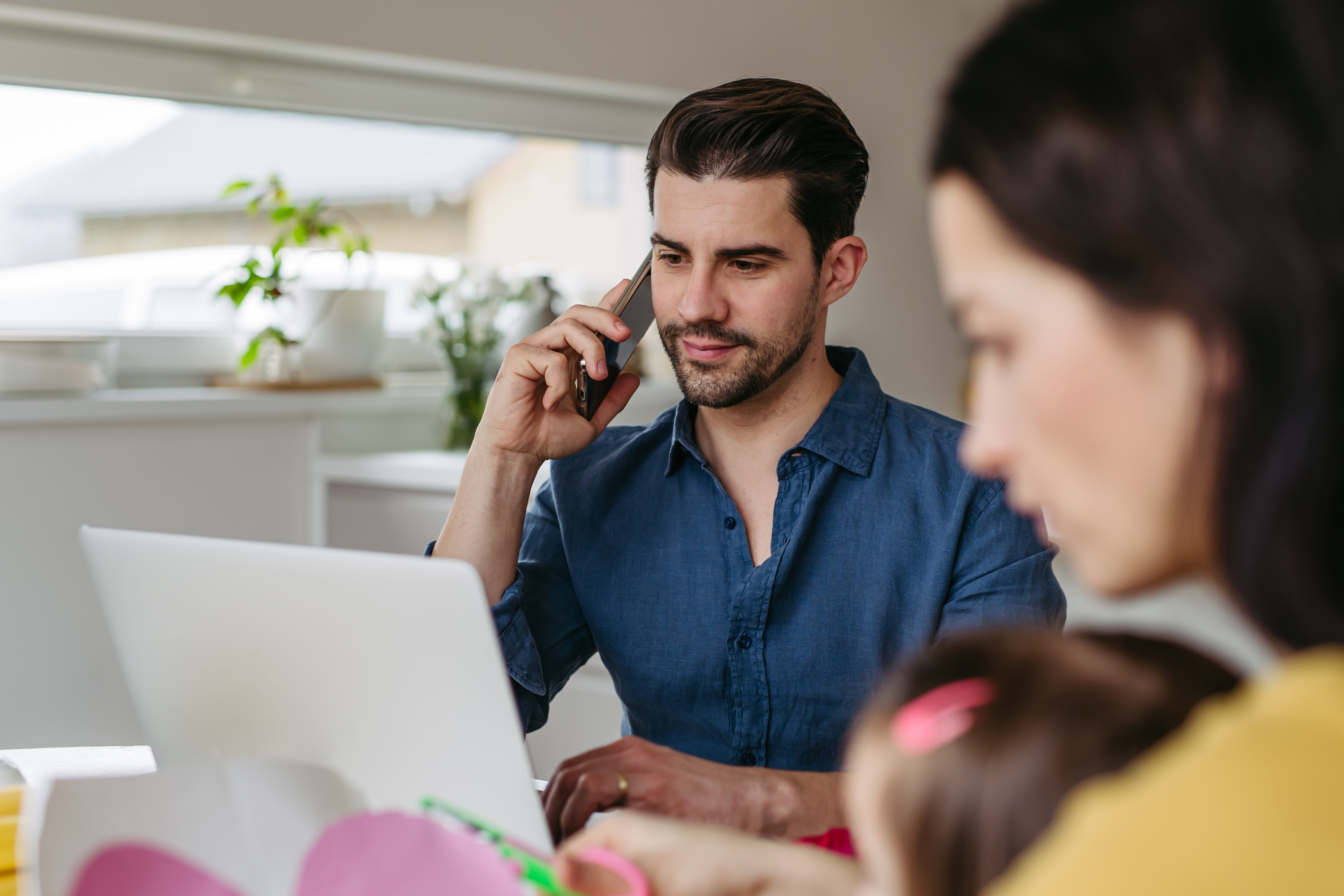 A man in a blue shirt works from home using a laptop and phone, while a woman, out of focus, tends to a child in the foreground