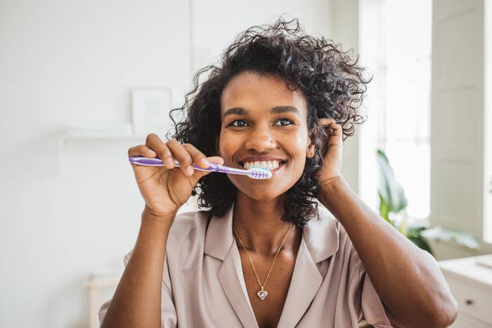 Person smiling while brushing teeth with a purple toothbrush in a bright, airy room