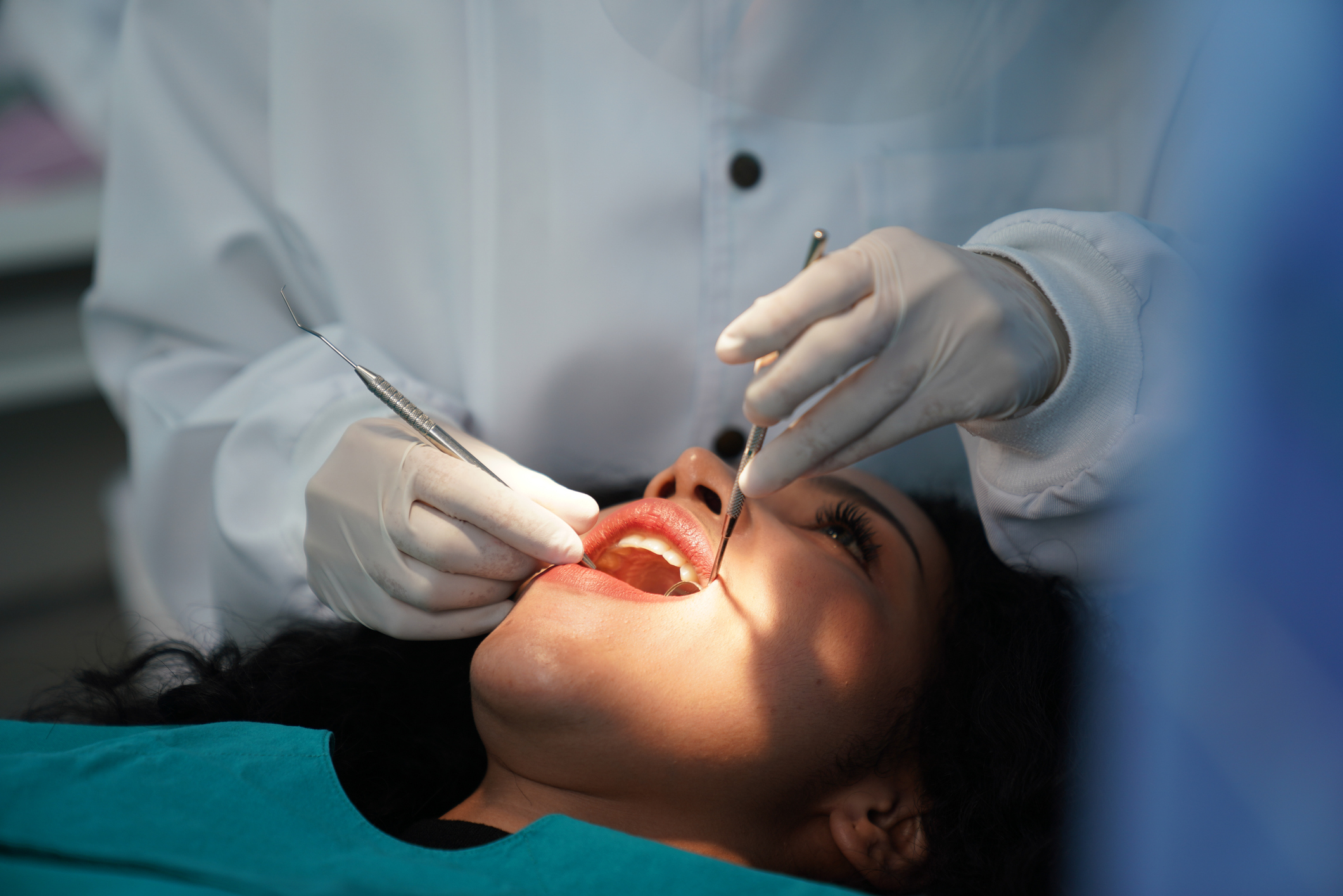 A dentist in a white coat and gloves performs a dental examination on a patient who is lying back in a dental chair, mouth open