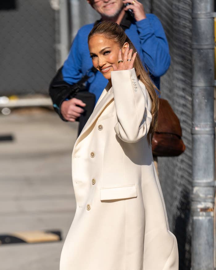 Jennifer Lopez waves to the camera, wearing a stylish long coat, while a man in the background talks on the phone