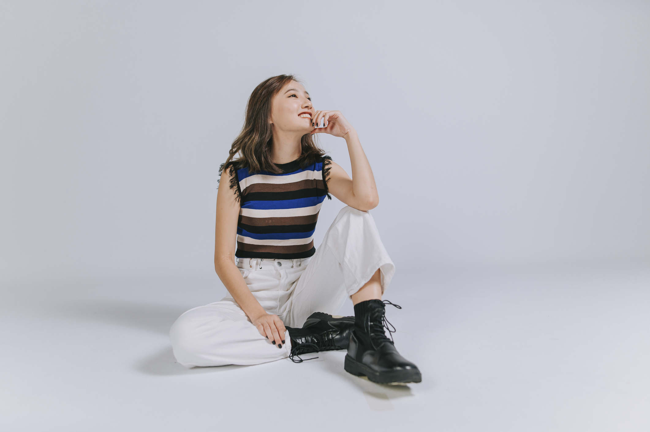 A woman in a striped top and white pants sits on the floor with one leg bent, smiling and resting her hand on her chin