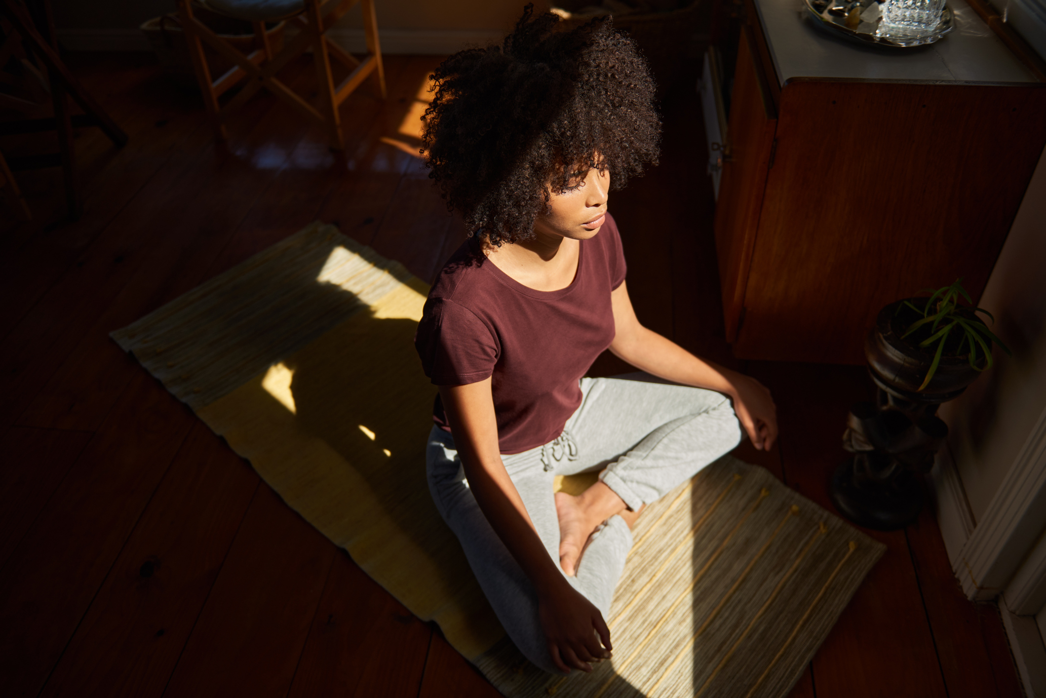 A person with curly hair sits cross-legged on a yoga mat in a peaceful indoor setting, engaging in a meditative practice