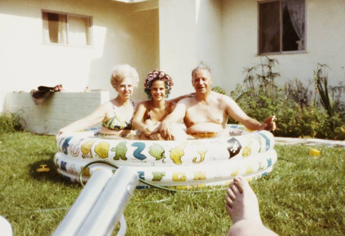 Three adults, two women and one man, sit in a kiddie pool in a backyard. One woman wears curlers. There are lawn and house surroundings visible