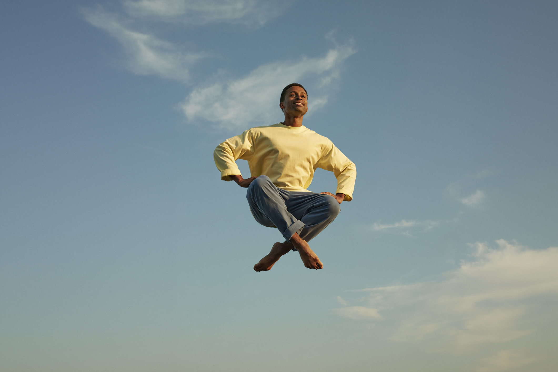 A person is mid-air in a meditation pose, smiling with arms bent and fists on hips, against a clear sky