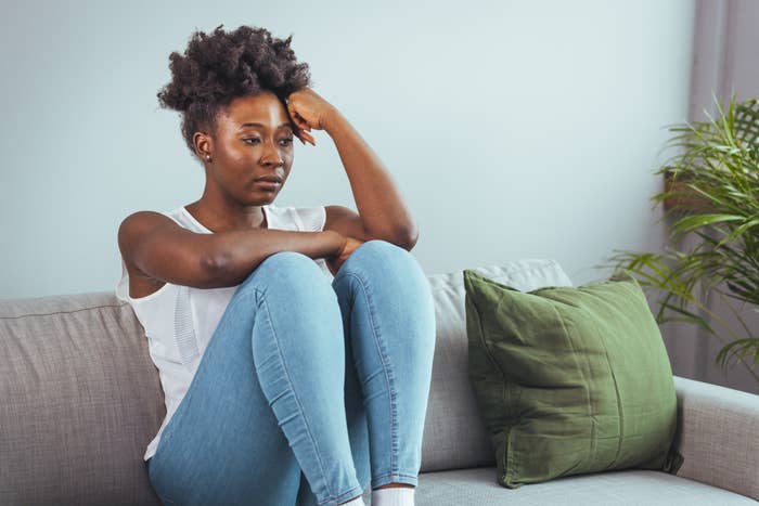 A woman sits on a couch wearing a white sleeveless shirt and jeans, looking contemplative with her head resting on her hand